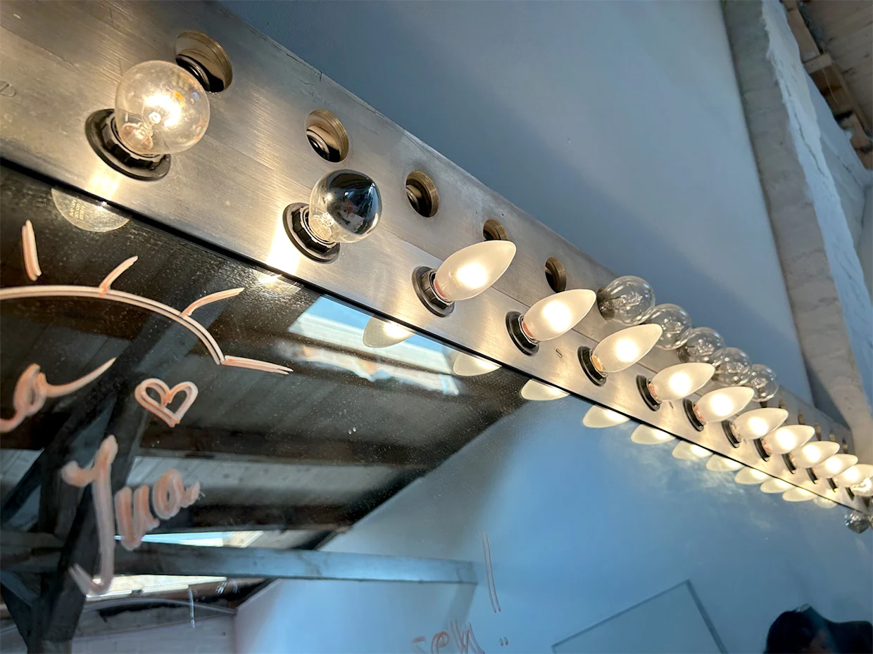 Spiegel mit einer Reihe von Glühbirnen, einige leuchten, andere sind ausgeschaltet, mit handgeschriebenen Zeichnungen und Text auf der Spiegeloberfläche. Spiegel mit einer Reihe von Glühbirnen, einige leuchten, andere sind ausgeschaltet, mit handgeschriebenen Zeichnungen und Text auf der Spiegeloberfläche.