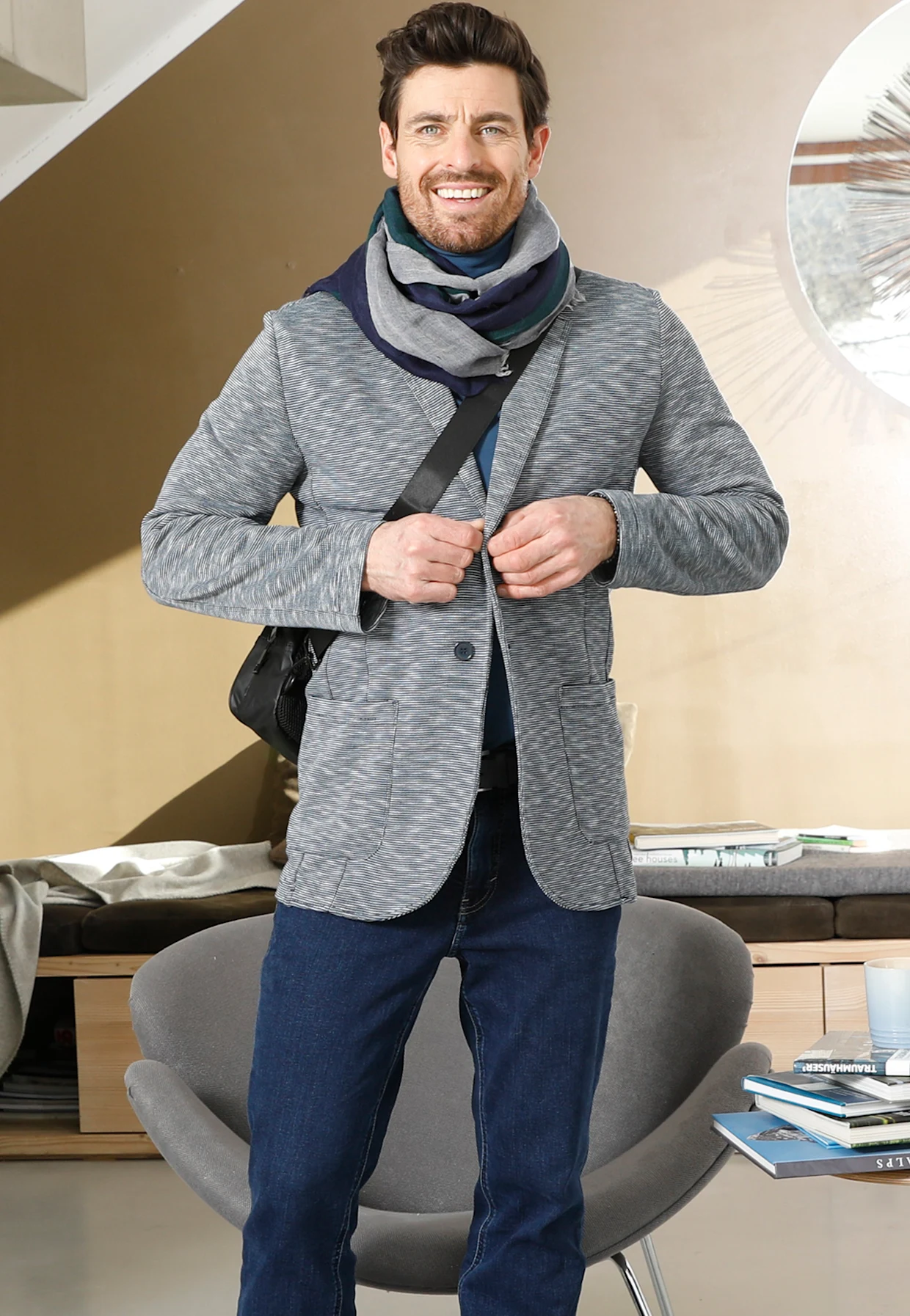 Mann trägt grauen Blazer, blauen Schal und Jeans, stehend in modernem Wohnzimmer mit grauem Sessel und Couchtisch mit Büchern. Mann trägt grauen Blazer, blauen Schal und Jeans, stehend in modernem Wohnzimmer mit grauem Sessel und Couchtisch mit Büchern.