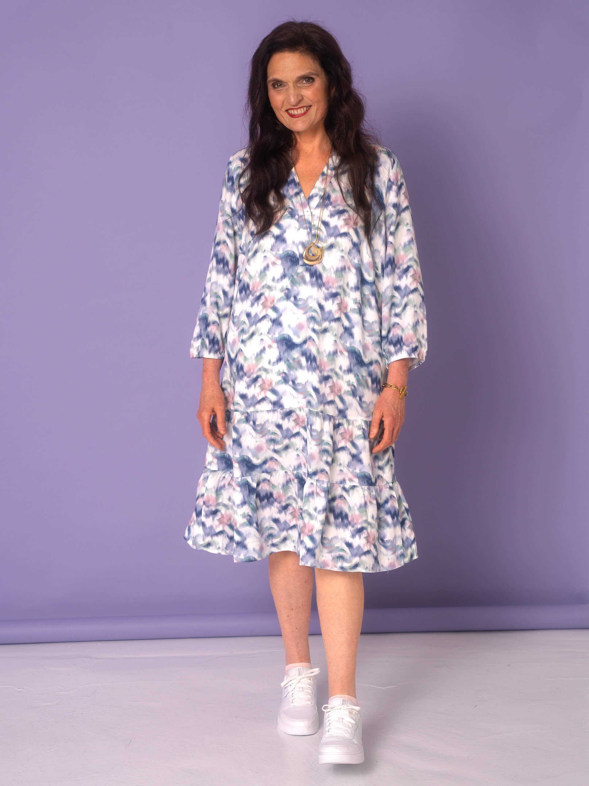 Ein Model trägt ein gemustertes Kleid mit langen Ärmeln und weiße Sneakers, während es vor einem lila Hintergrund steht.