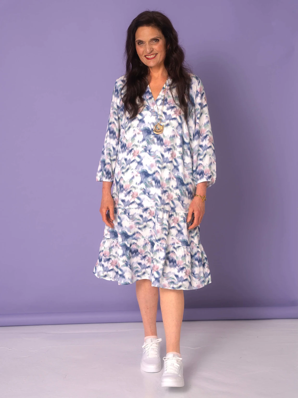 Ein Model trägt ein gemustertes Kleid mit langen Ärmeln und weiße Sneakers, während es vor einem lila Hintergrund steht.