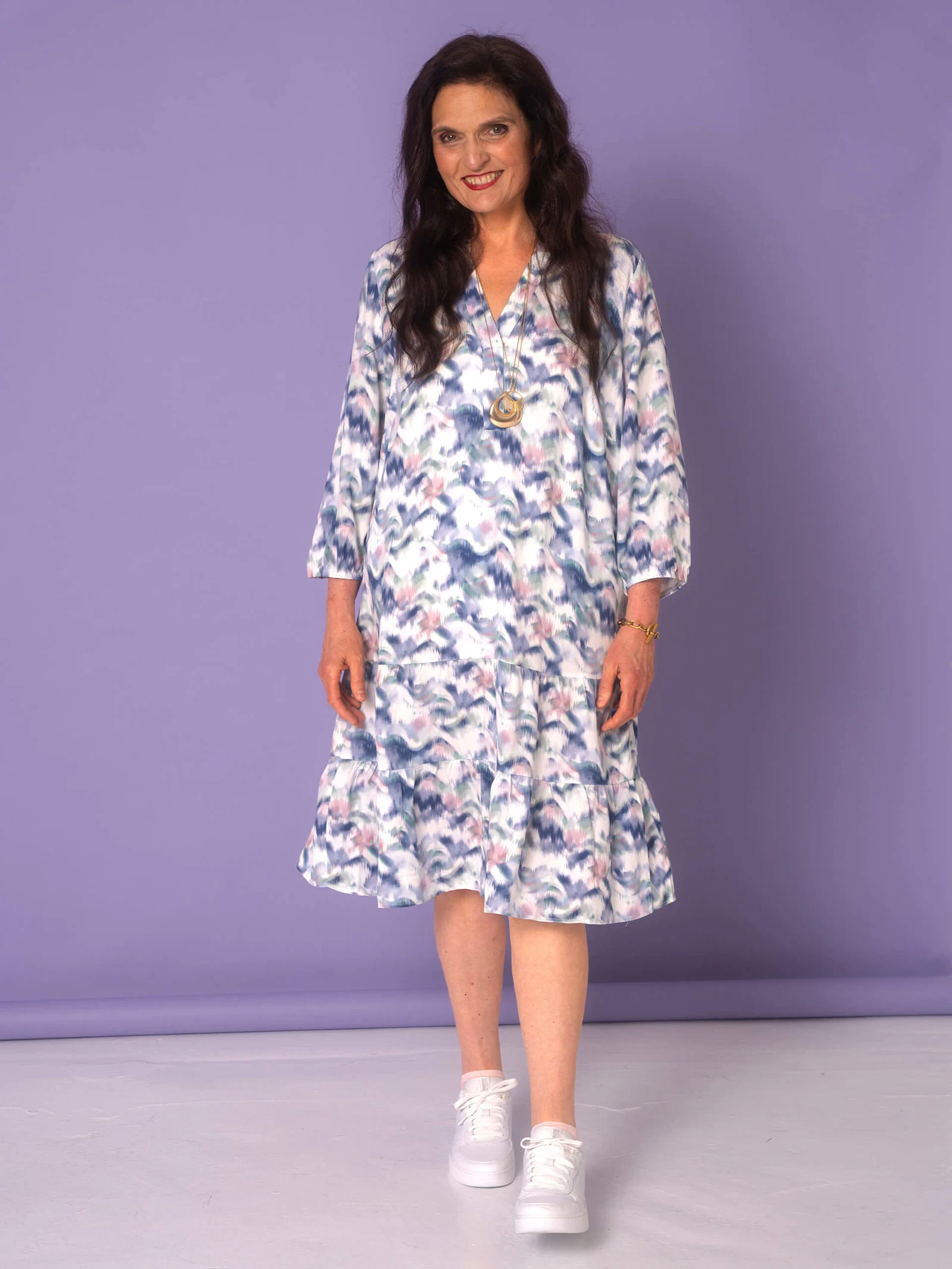 Ein Model trägt ein gemustertes Kleid mit langen Ärmeln und weiße Sneakers, während es vor einem lila Hintergrund steht.