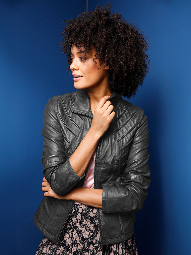 Ein Model trägt eine schwarze, gesteppte Lederjacke mit Stehkragen, kombiniert mit einem gemusterten Rock, vor blauem Hintergrund.