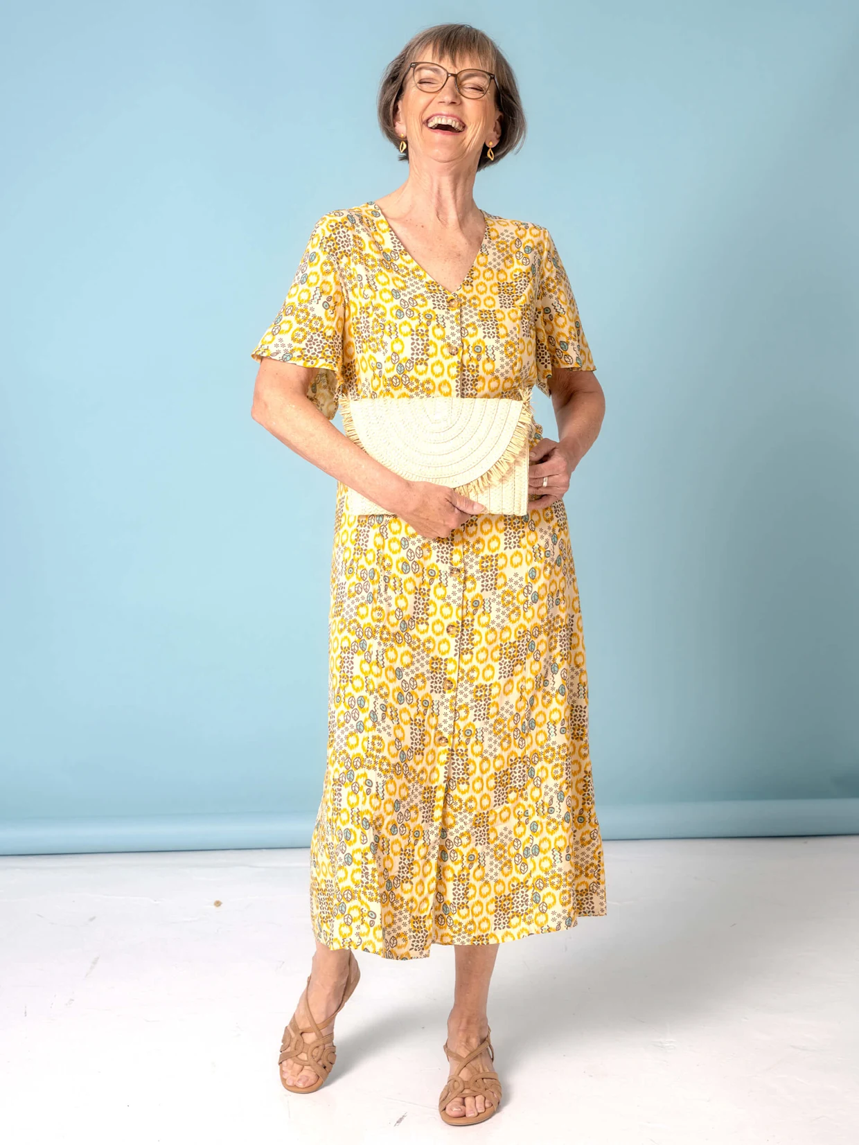 Eine Frau trägt ein gelbes, gemustertes Kleid mit kurzen Ärmeln und hält eine helle Clutch. Dazu trägt sie Sandalen. Der Hintergrund ist hellblau.