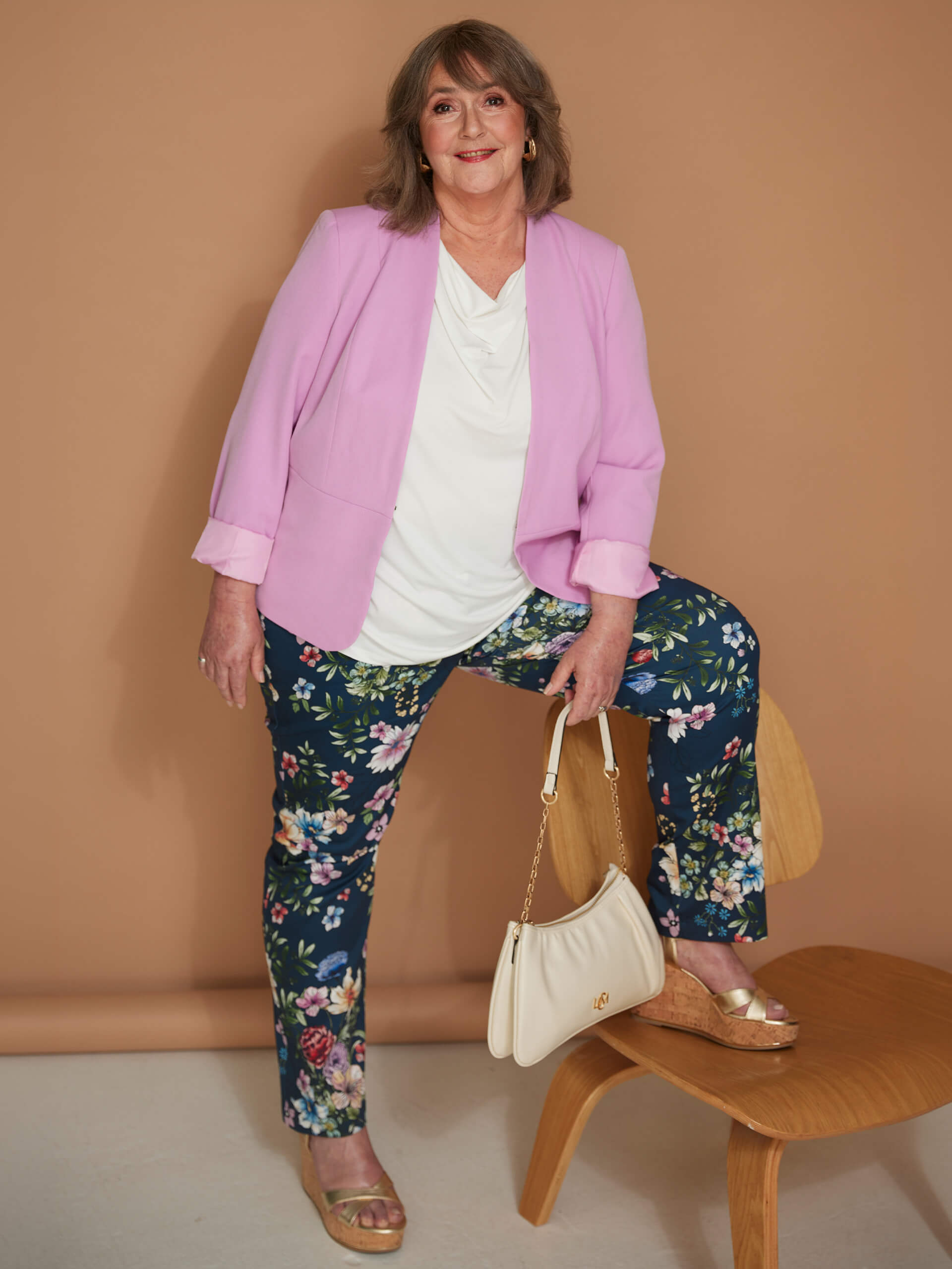 Ein Model trägt rosa Blazer, weißes Shirt und geblümte Hose, mit einem Fuß auf einem Holzstuhl vor braunem Hintergrund.