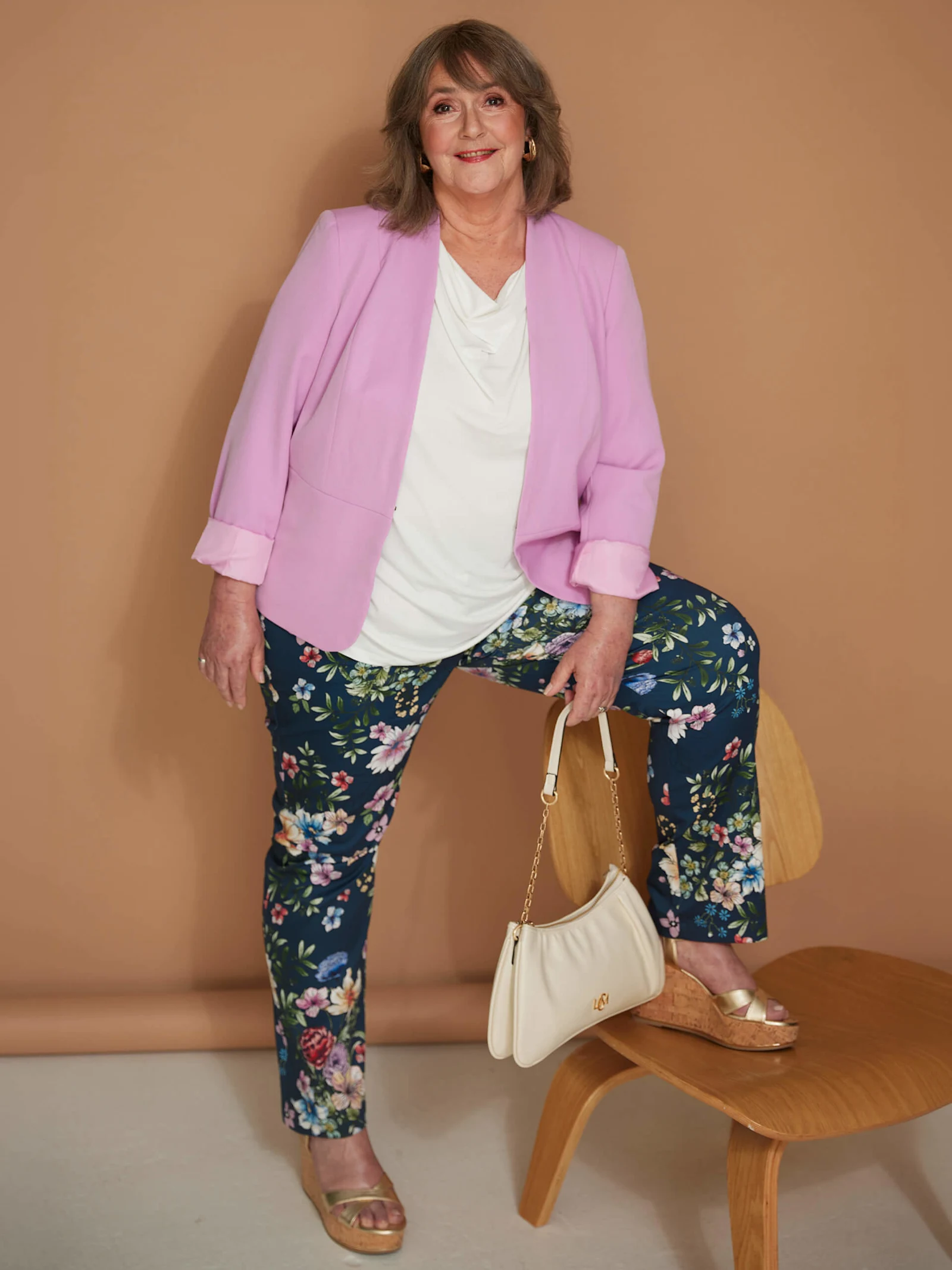 Ein Model trägt rosa Blazer, weißes Shirt und geblümte Hose, mit einem Fuß auf einem Holzstuhl vor braunem Hintergrund.