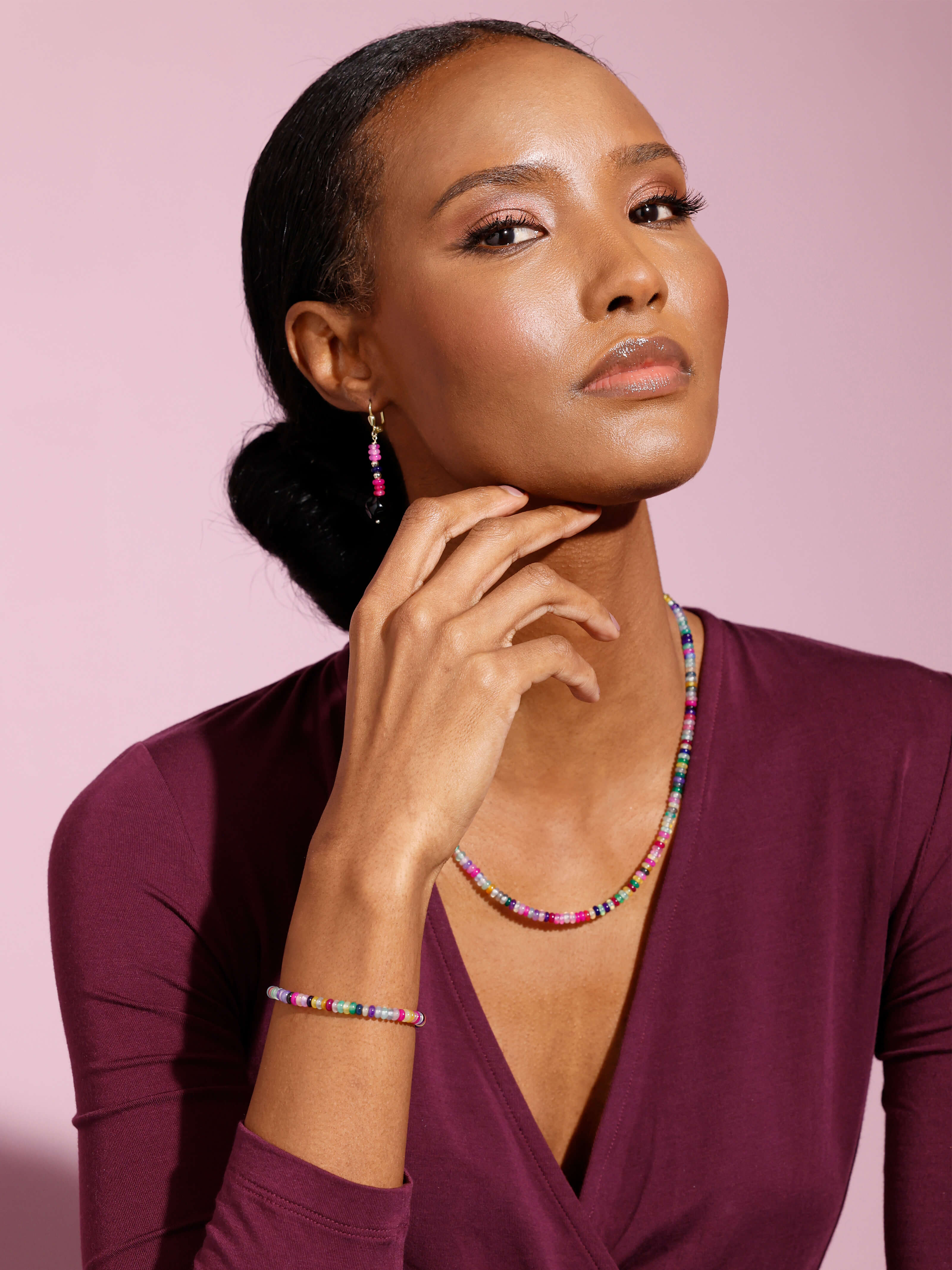 Ein Model trägt ein bordeauxfarbenes Oberteil und bunten Schmuck, sitzt vor pastellfarbenem Hintergrund.
