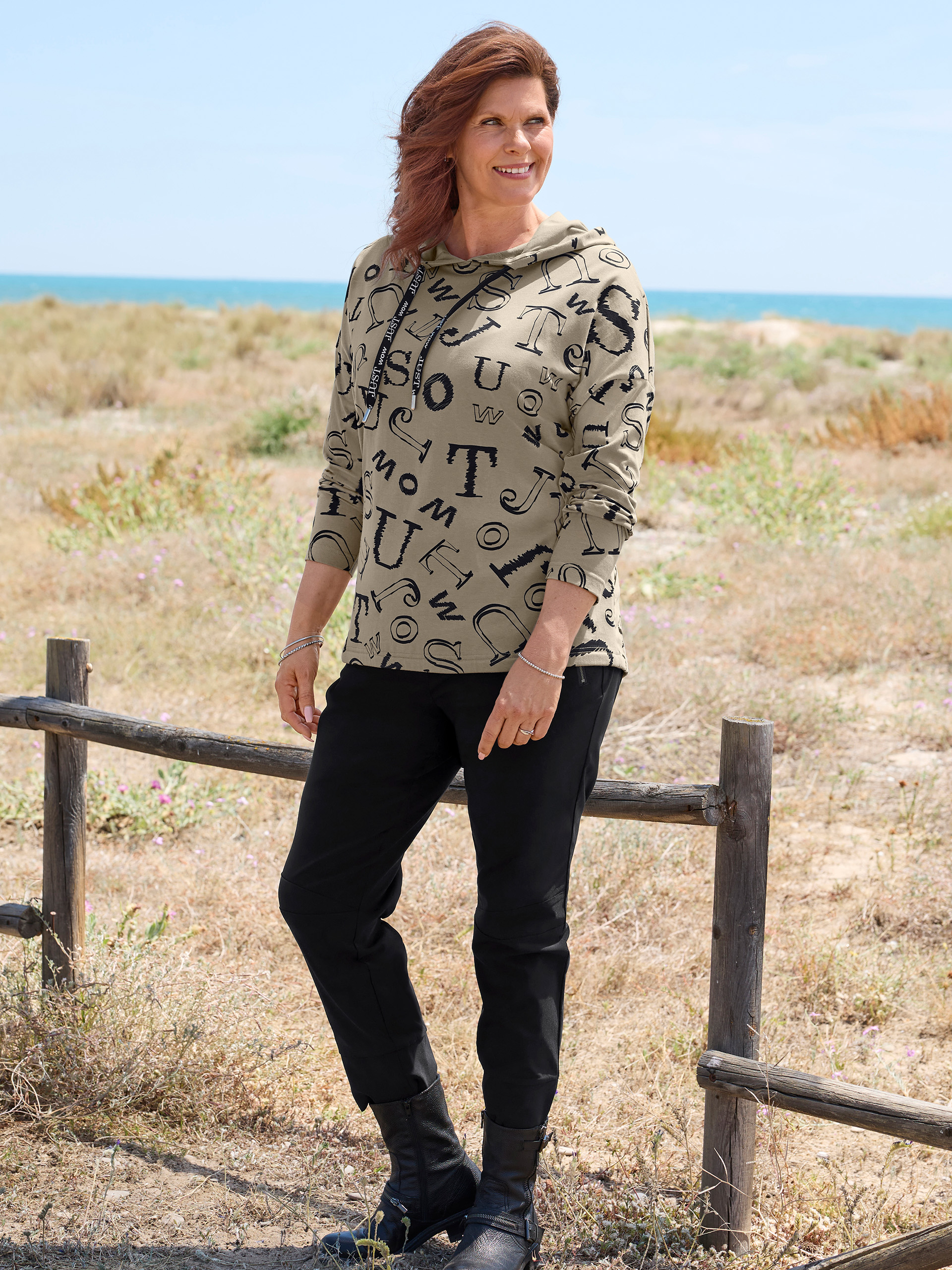Een glimlachende vrouw met roodbruin haar staat op een strandpad. Ze draagt een kaki hoodie met zwarte letters, een zwarte broek en zwarte laarzen. Op de achtergrond zijn de zee en een houten hek te zien.