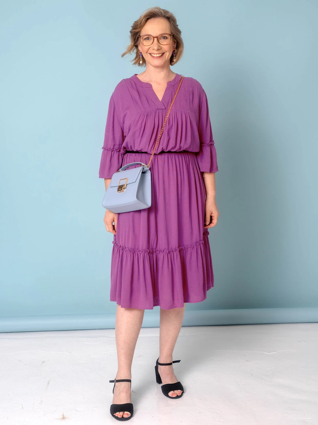 Eine Frau trägt ein lila Kleid mit Rüschendetails und eine kleine, hellblaue Tasche.