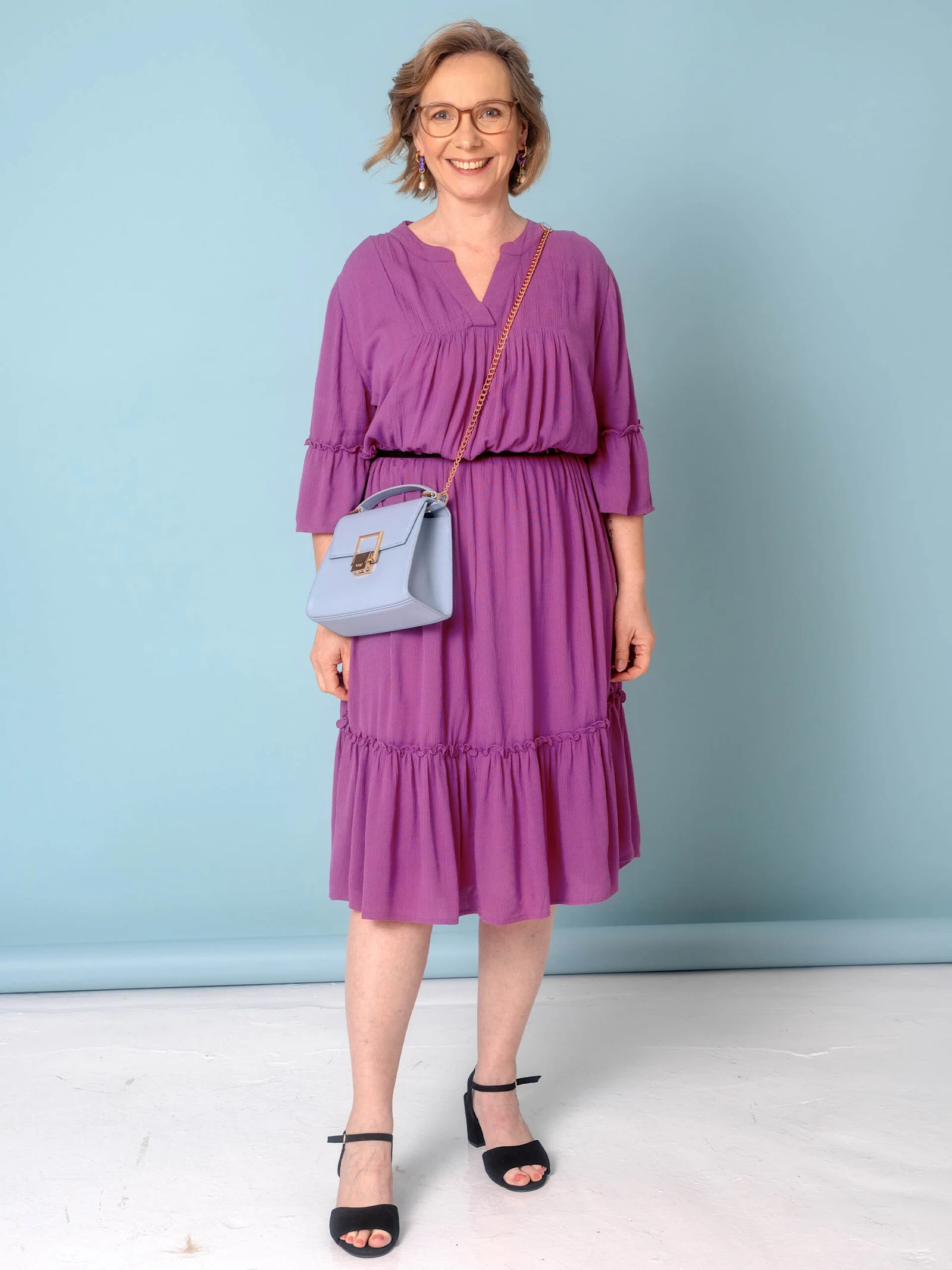 Eine Frau trägt ein lila Kleid mit Rüschendetails und eine kleine, hellblaue Tasche.
