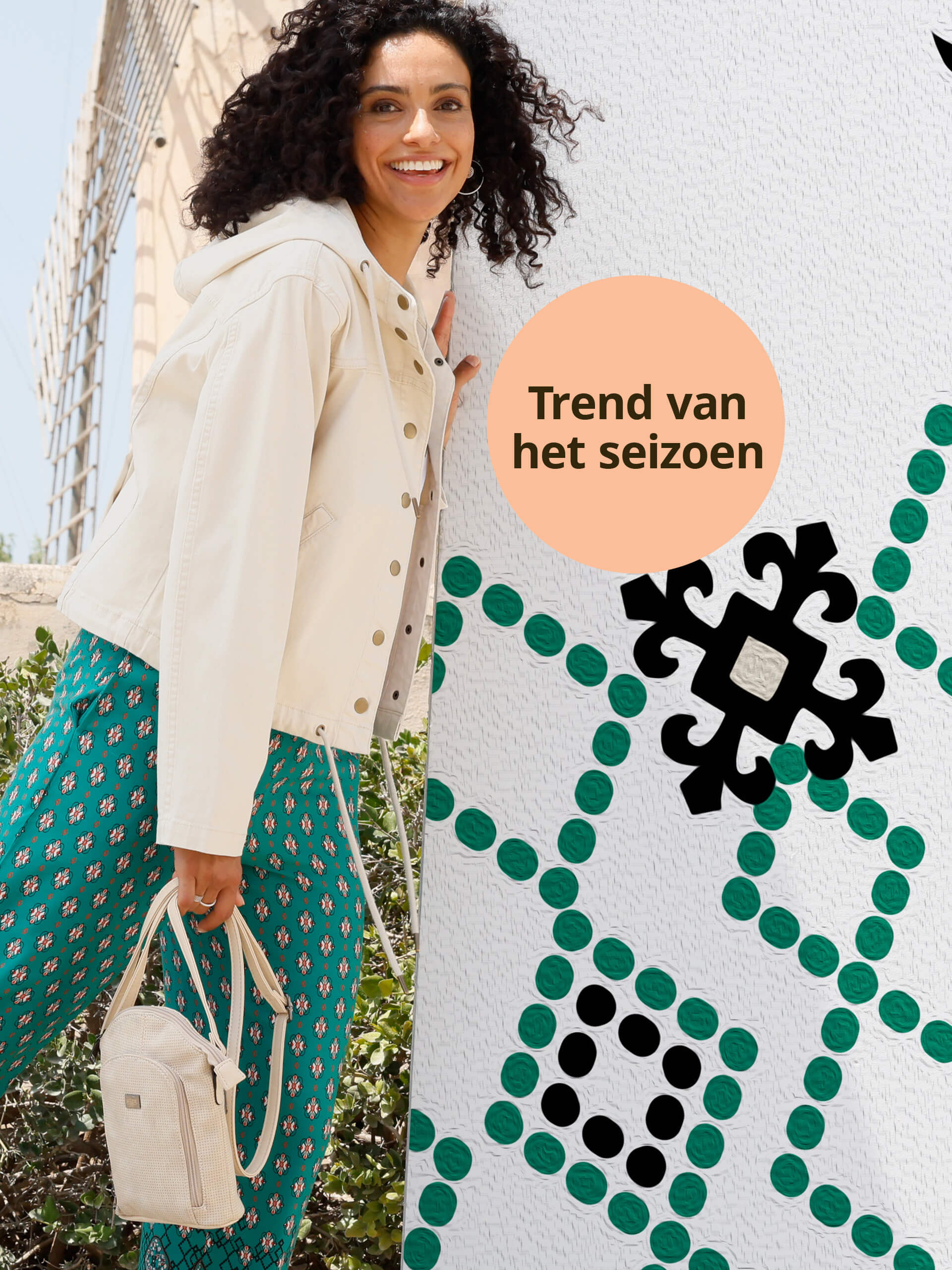Een model draagt een crèmekleurig jasje en een groene broek met patroon, met daarnaast een oranje cirkel met de tekst 'Trend van het seizoen'.