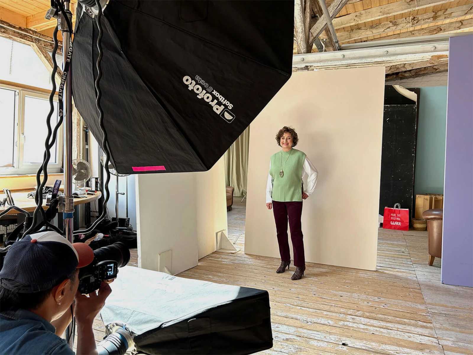 Ein Model steht vor einem beigen Hintergrund in einem Fotostudio, trägt eine grüne Strickweste, weiße Bluse, bordeauxrote Hose und Leo-Stiefeletten, Fotograf mit Kamera und Lichttechnik sichtbar. Ein Model steht vor einem beigen Hintergrund in einem Fotostudio, trägt eine grüne Strickweste, weiße Bluse, bordeauxrote Hose und Leo-Stiefeletten, Fotograf mit Kamera und Lichttechnik sichtbar.
