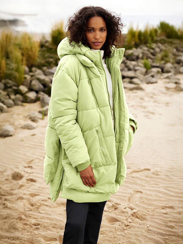 Ein Model trägt eine pistaziefarbene, dicke Steppjacke mit Kapuze, kombiniert mit schwarzen Hosen, und steht am Strand.