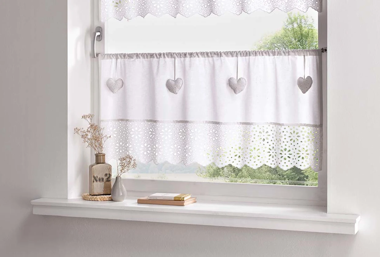 Weiße Fensterdeko mit Herzanhängern und Spitzenmuster, auf einer Fensterbank stehen dekorative Vasen. Weiße Fensterdeko mit Herzanhängern und Spitzenmuster, auf einer Fensterbank stehen dekorative Vasen.