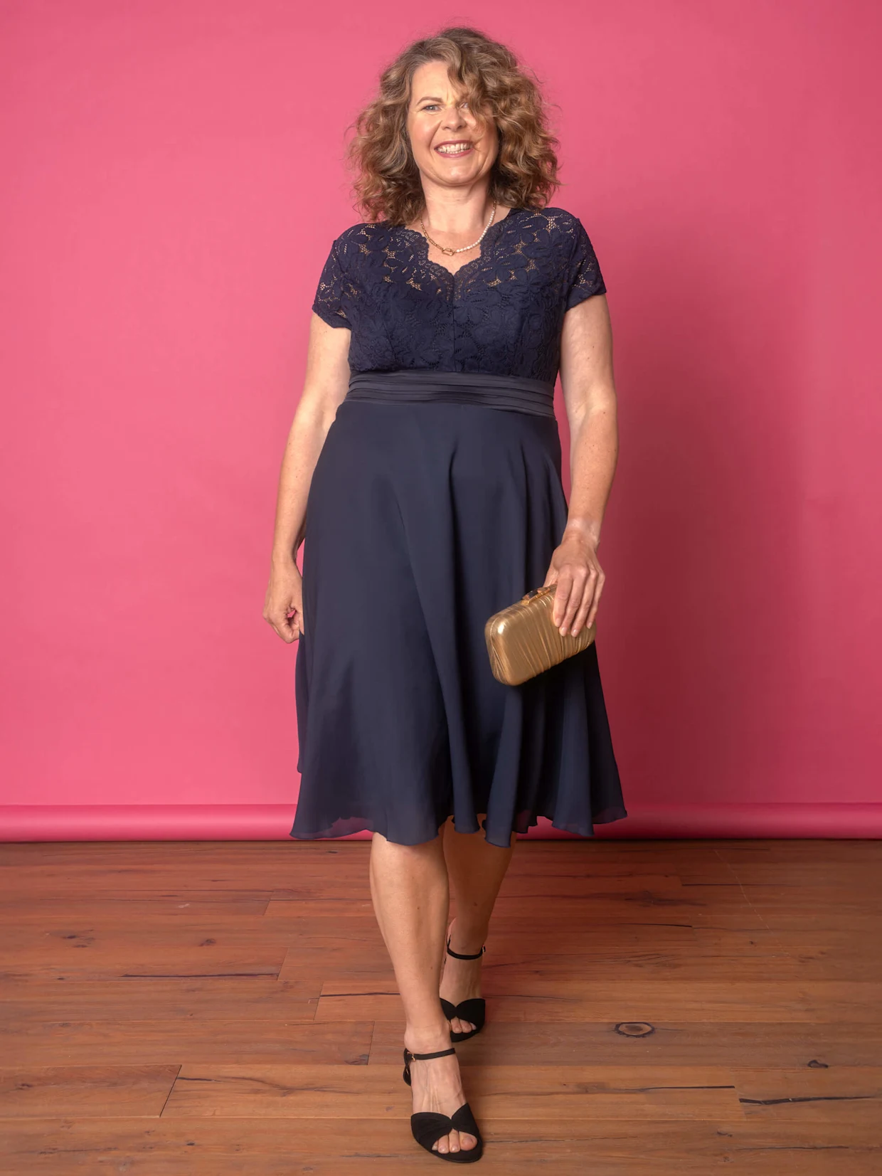 Eine Frau trägt ein dunkelblaues Kleid mit Spitzenoberteil und hält eine goldene Clutch, während sie vor einem pinken Hintergrund steht.