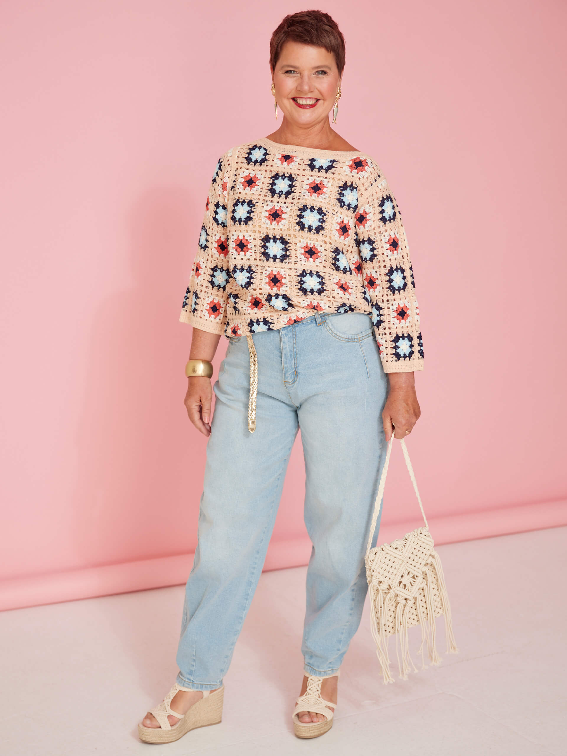 Ein Model trägt einen gehäkelten Pullover mit bunten Quadraten, helle Jeans, Sandaletten mit Keilabsatz und hält eine gehäkelte Tasche vor rosa Hintergrund.