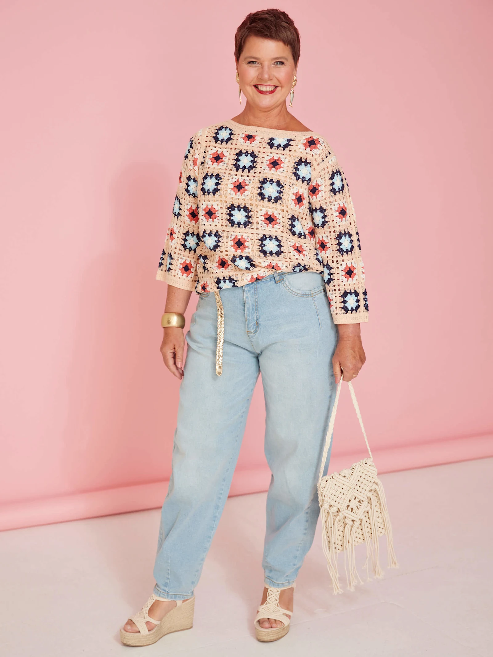 Ein Model trägt einen gehäkelten Pullover mit bunten Quadraten, helle Jeans, Sandaletten mit Keilabsatz und hält eine gehäkelte Tasche vor rosa Hintergrund.
