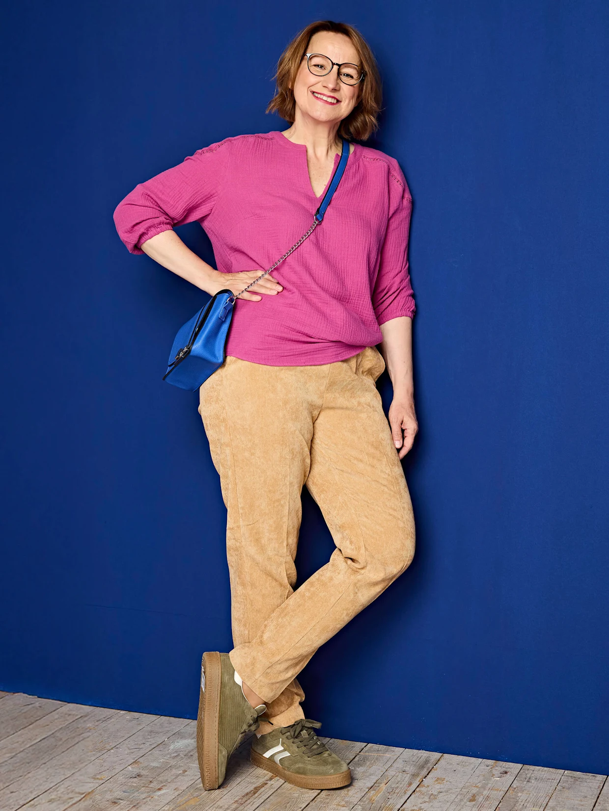 Eine Frau trägt eine pinke Bluse mit 3/4-Ärmeln, eine beige Cordhose, olivgrüne Sneaker und eine blaue Umhängetasche vor blauem Hintergrund.