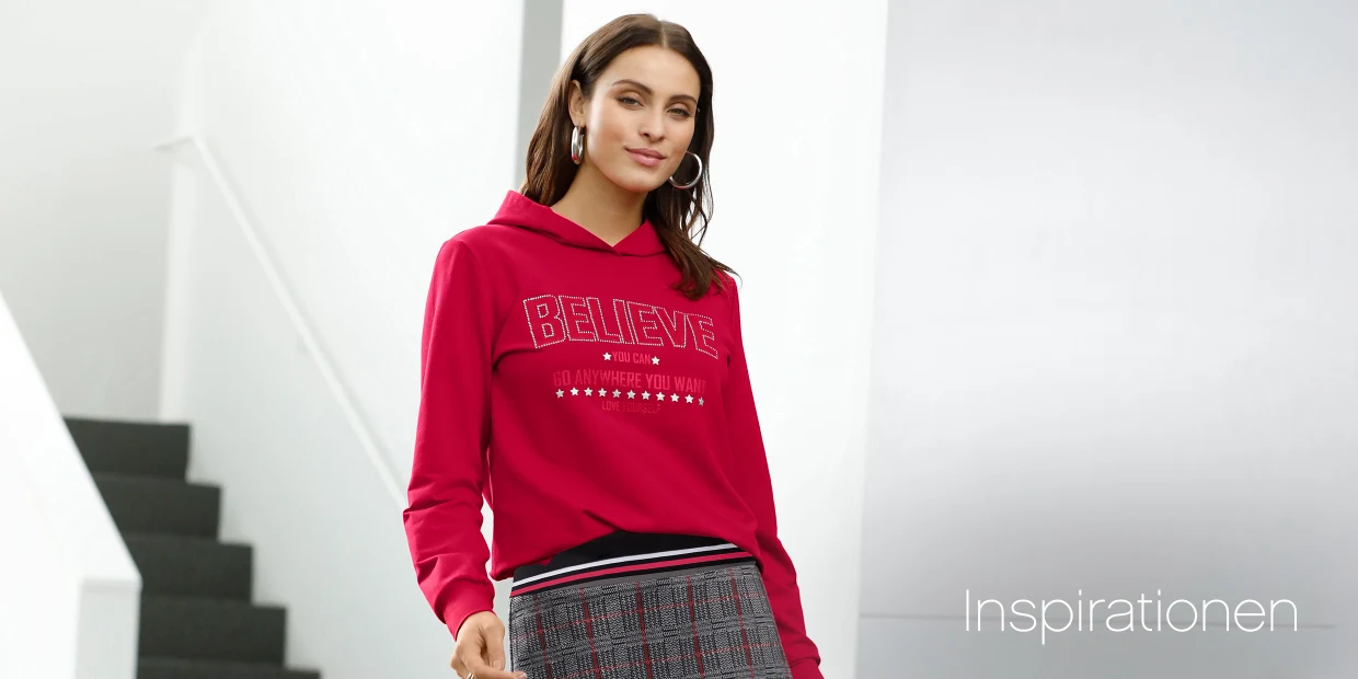 Frau trägt roten Hoodie mit Schriftzug "BELIEVE" und grauen Rock mit Karomuster vor modernem Hintergrund mit Treppe. Frau trägt roten Hoodie mit Schriftzug "BELIEVE" und grauen Rock mit Karomuster vor modernem Hintergrund mit Treppe.