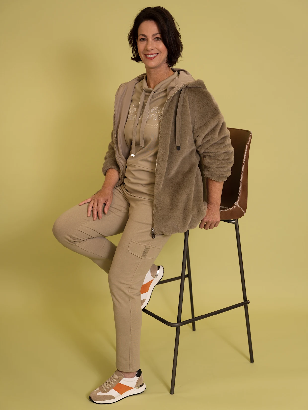 Eine dunkelhaarige Frau trägt eine schlammfarbene Fellimitatjacke mit Gummizug an Ärmeln und Saum, ein schlammfarbenes Stehkragenshirt mit Glitzersteinchen, schlammfarbene Jogg Pants und weiß-orange Sneakers. Der Hintergrund ist gelb-golden.