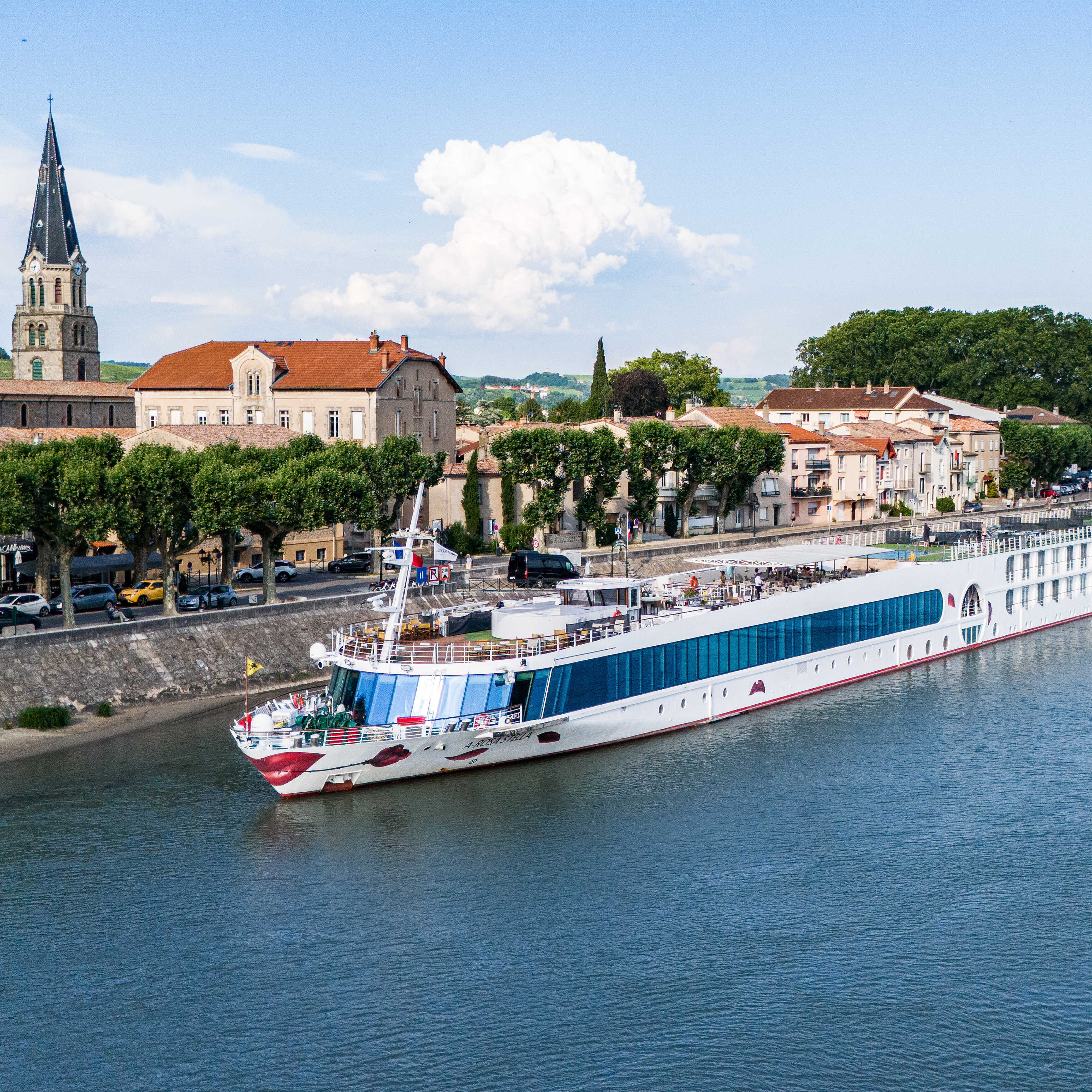 Großes Flusskreuzfahrtschiff fährt an einer Stadt mit Kirche und Bäumen am Ufer vorbei. Blauer Himmel mit Wolken.

