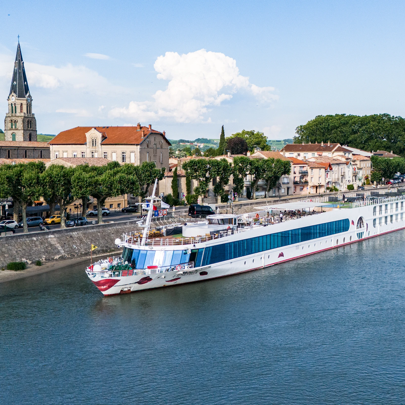 Großes Flusskreuzfahrtschiff fährt an einer Stadt mit Kirche und Bäumen am Ufer vorbei. Blauer Himmel mit Wolken.
Großes Flusskreuzfahrtschiff fährt an einer Stadt mit Kirche und Bäumen am Ufer vorbei. Blauer Himmel mit Wolken.