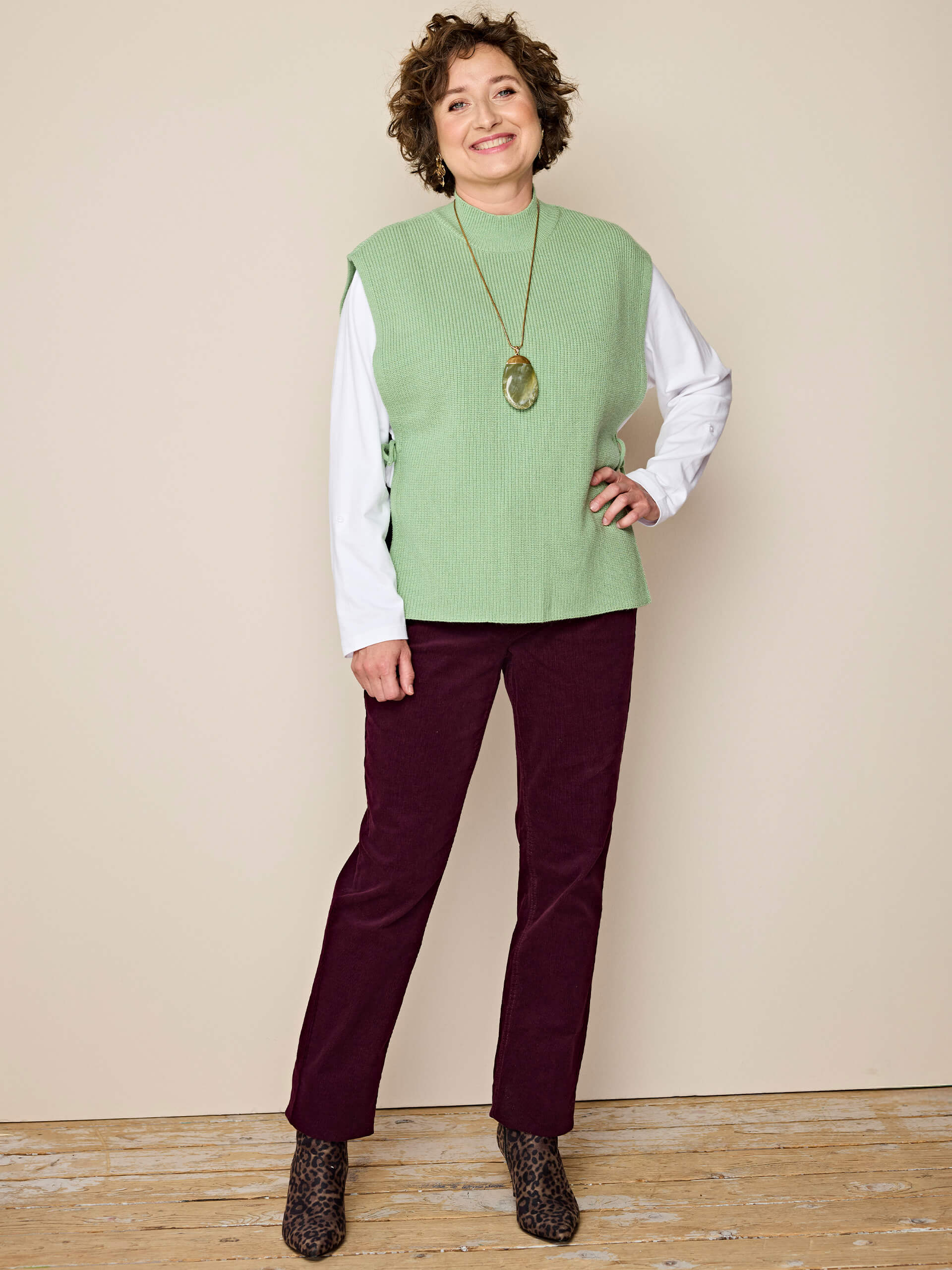 Ein Model trägt eine grüne Strickweste über weißem Langarmshirt, bordeauxrote Cordhose und Leo-Stiefeletten, vor beigem Hintergrund.