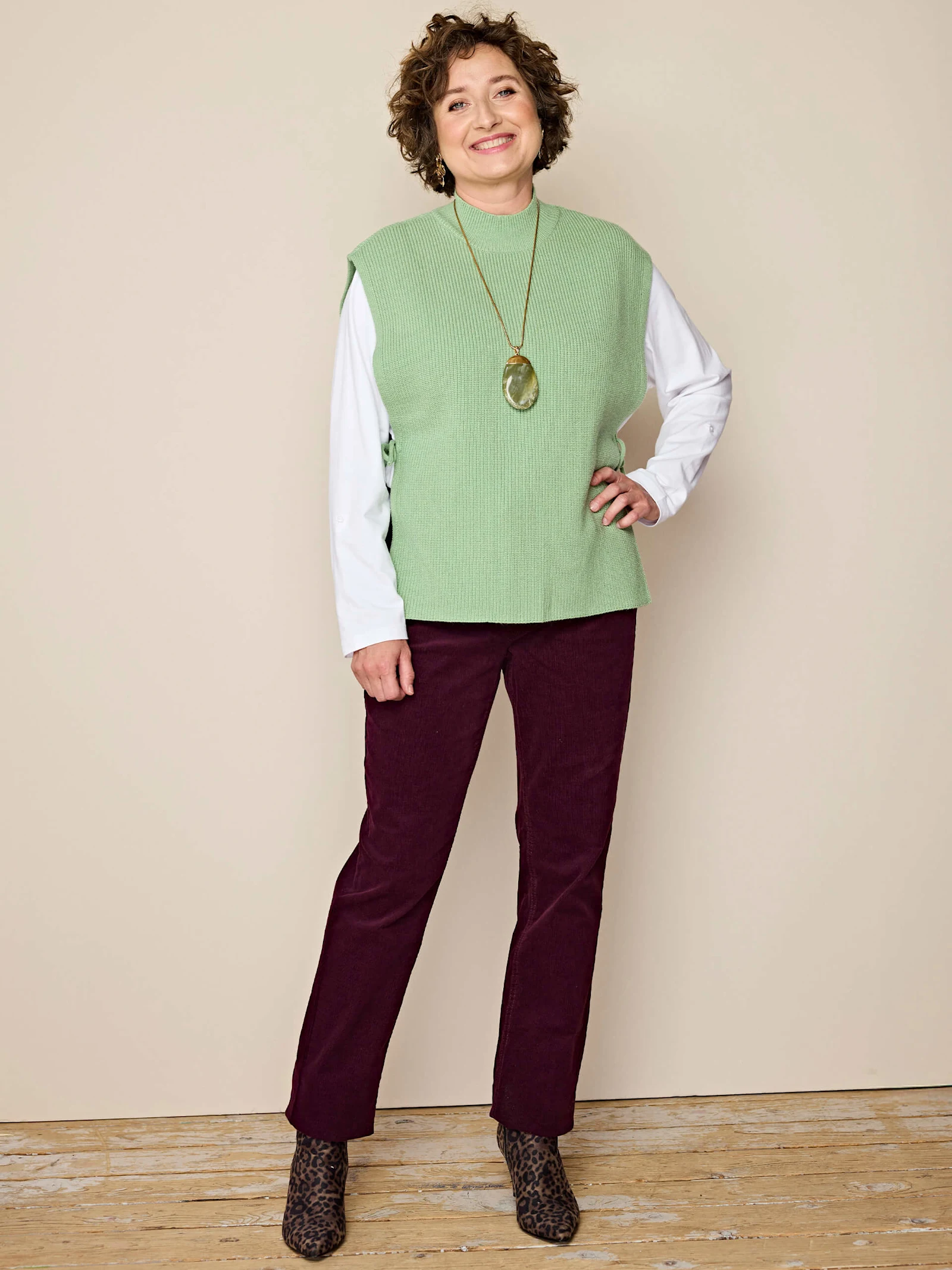 Ein Model trägt eine grüne Strickweste über weißem Langarmshirt, bordeauxrote Cordhose und Leo-Stiefeletten, vor beigem Hintergrund. Ein Model trägt eine grüne Strickweste über weißem Langarmshirt, bordeauxrote Cordhose und Leo-Stiefeletten, vor beigem Hintergrund.