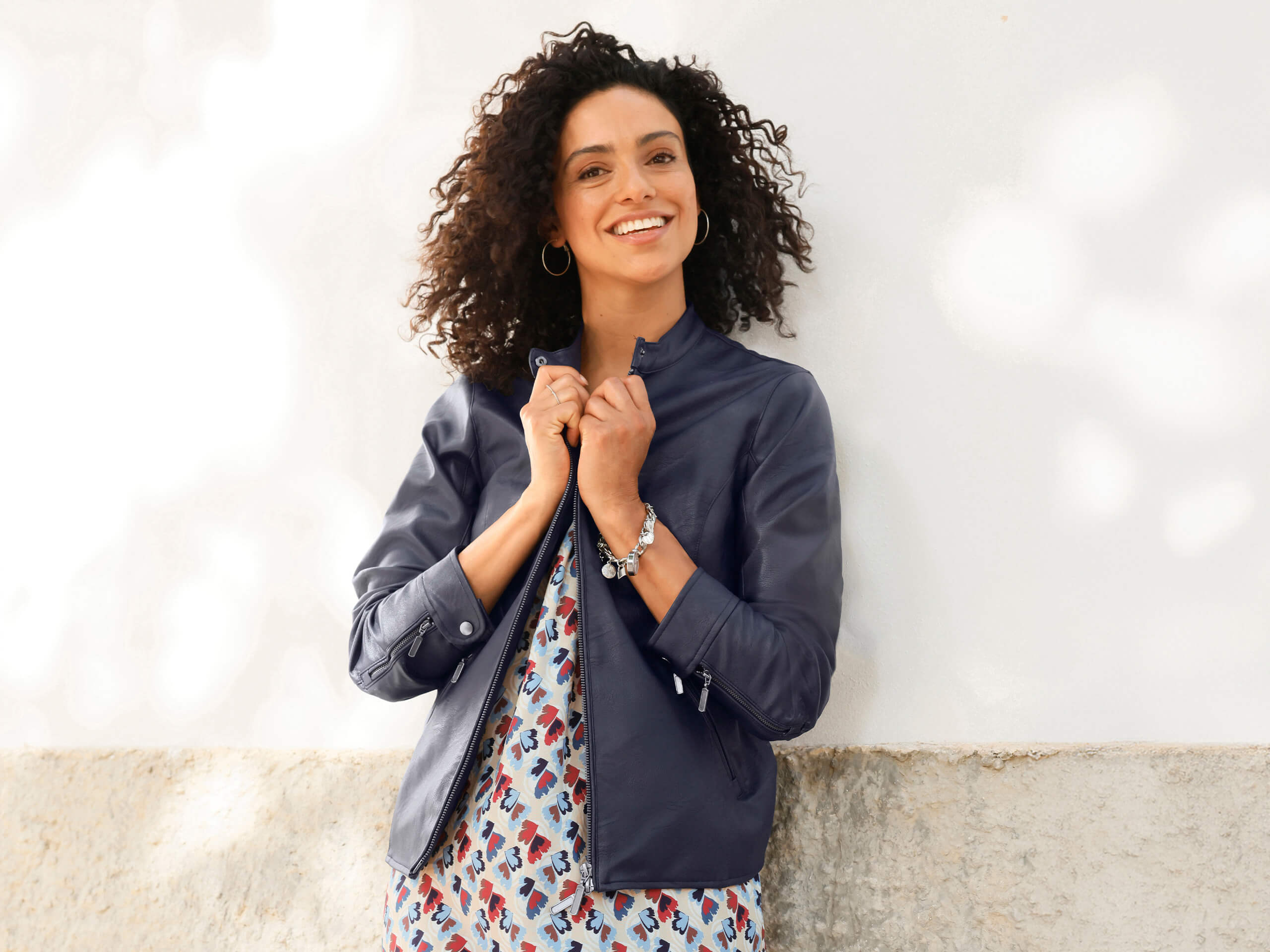 Ein Model trägt eine dunkelblaue Lederjacke über einem gemusterten Kleid mit abstraktem Design, steht vor einer hellen Wand.