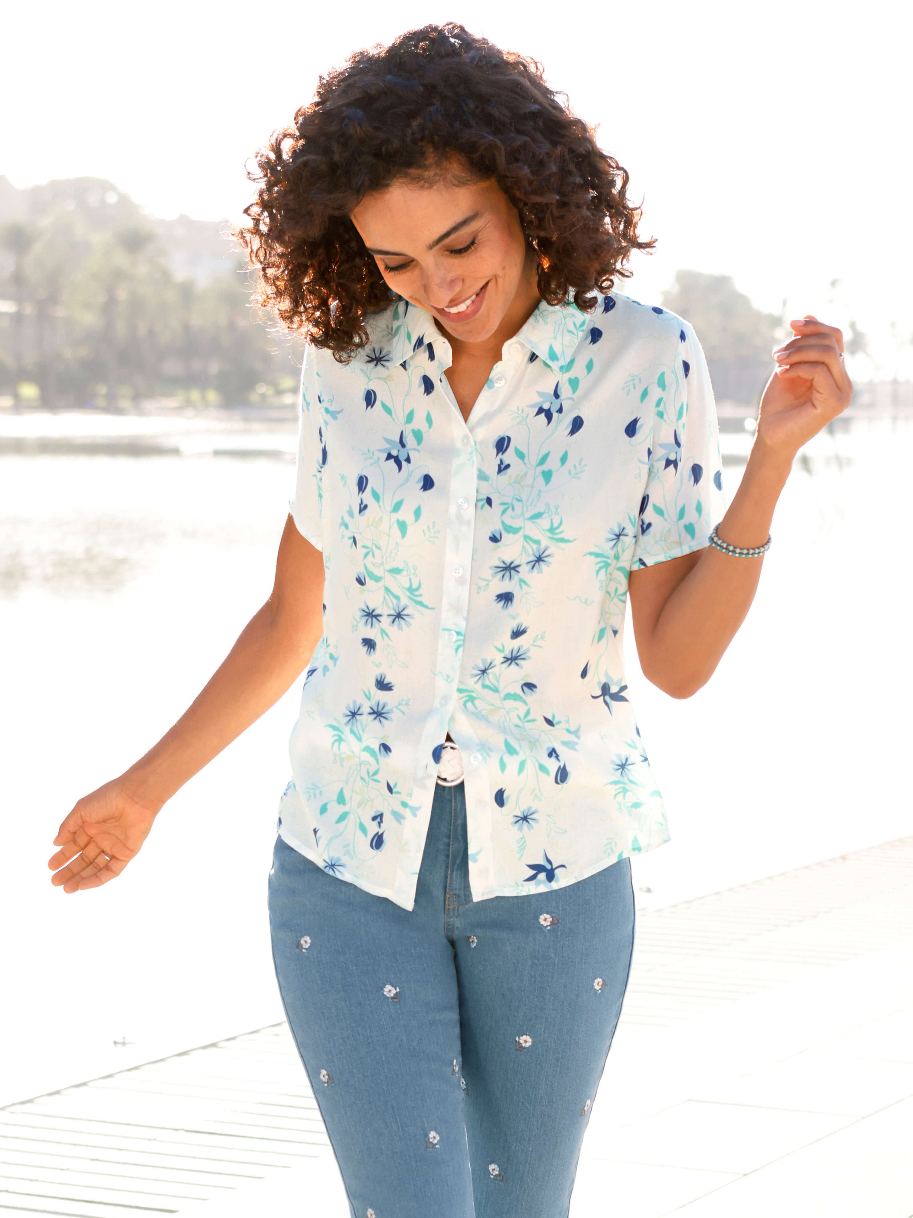 Ein Model trägt eine helle, kurzärmelige Bluse mit blau-grünem Blumenmuster und eine hellblaue bestickte Jeans, steht am Wasser.