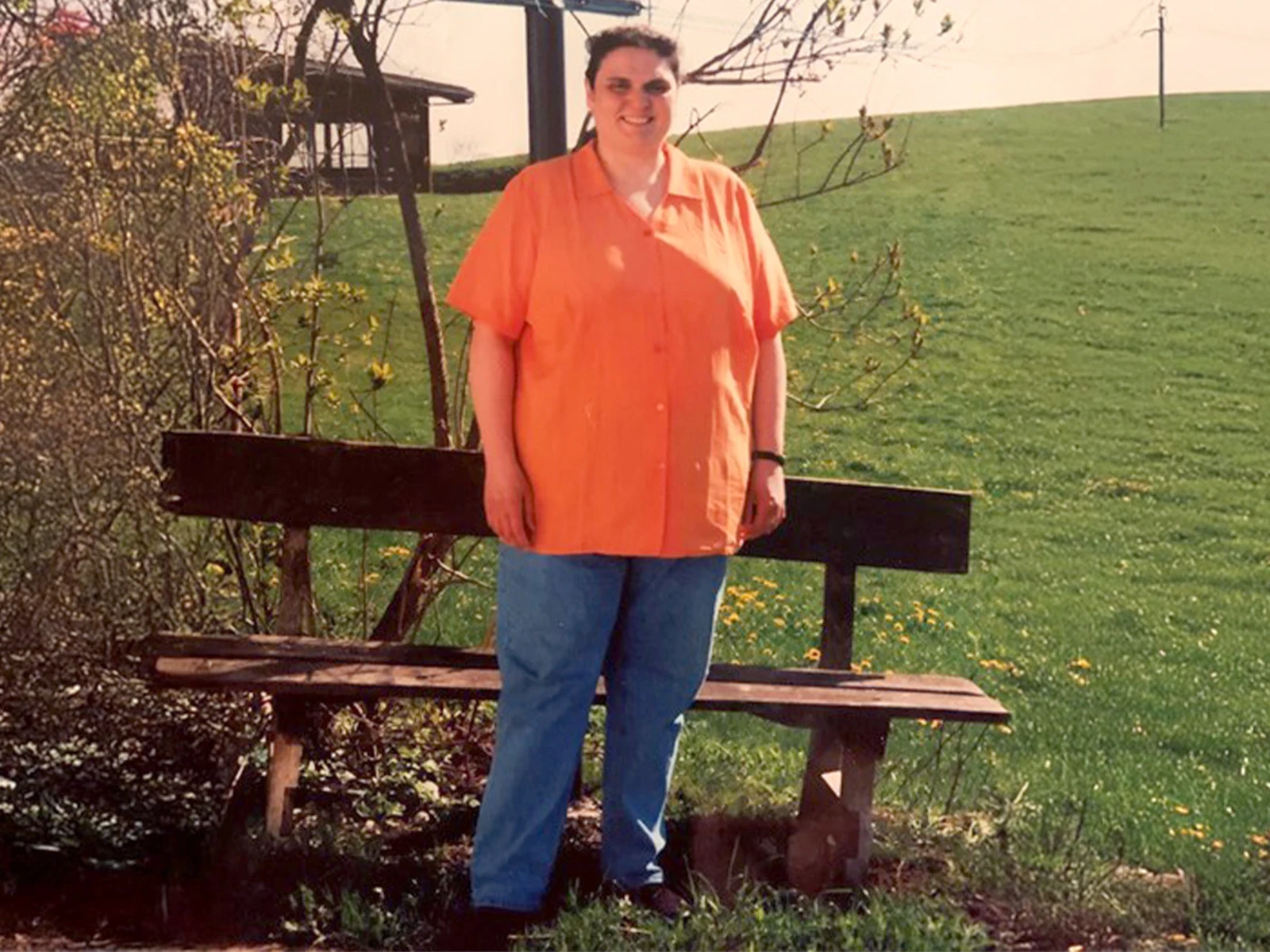 Eine Frau steht vor einer Bank im Freien, trägt ein orangefarbenes Hemd und eine blaue Jeans. Im Hintergrund sind eine grüne Wiese und Bäume sichtbar. Eine Frau steht vor einer Bank im Freien, trägt ein orangefarbenes Hemd und eine blaue Jeans. Im Hintergrund sind eine grüne Wiese und Bäume sichtbar.