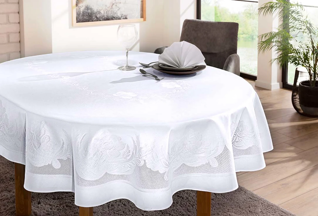 Runder Esstisch mit weißer, floraler Tischdecke, gedeckt mit Teller, Besteck, gefalteter Serviette und Weinglas in einem hellen Wohnraum mit Pflanzen. Runder Esstisch mit weißer, floraler Tischdecke, gedeckt mit Teller, Besteck, gefalteter Serviette und Weinglas in einem hellen Wohnraum mit Pflanzen.
