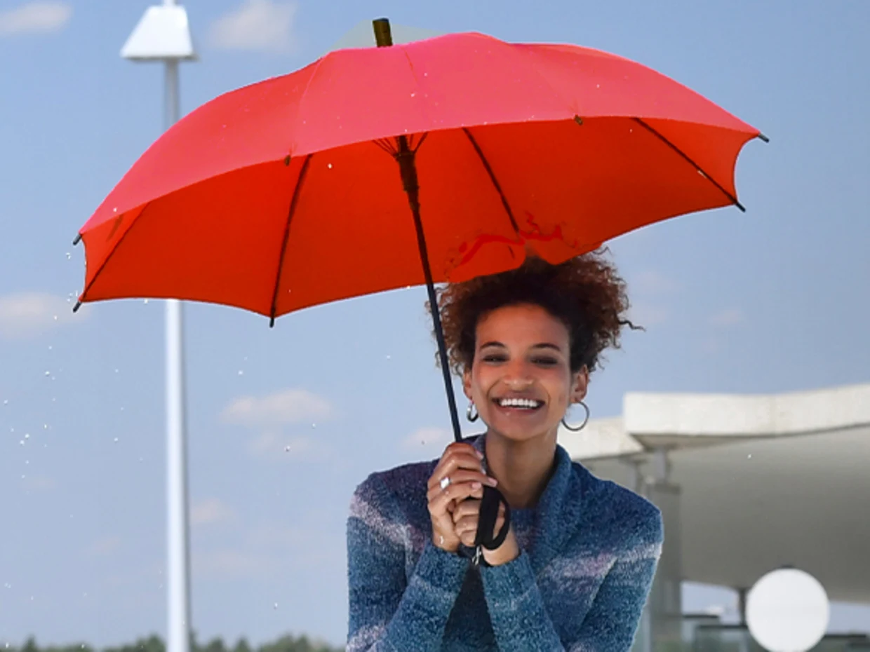 Eine Frau hält einen roten Regenschirm vor einem blauen Himmel mit vereinzelten Wolken.