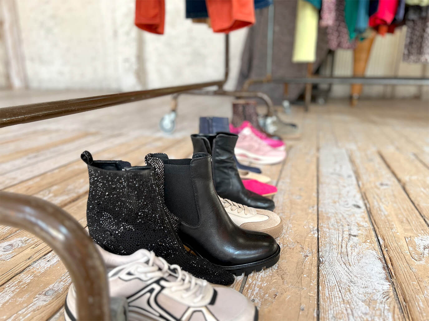 Mehrere Paar Schuhe, darunter schwarze Stiefeletten und helle Sneaker, stehen auf einem Holzboden vor einer Kleiderstange mit bunten Kleidungsstücken.