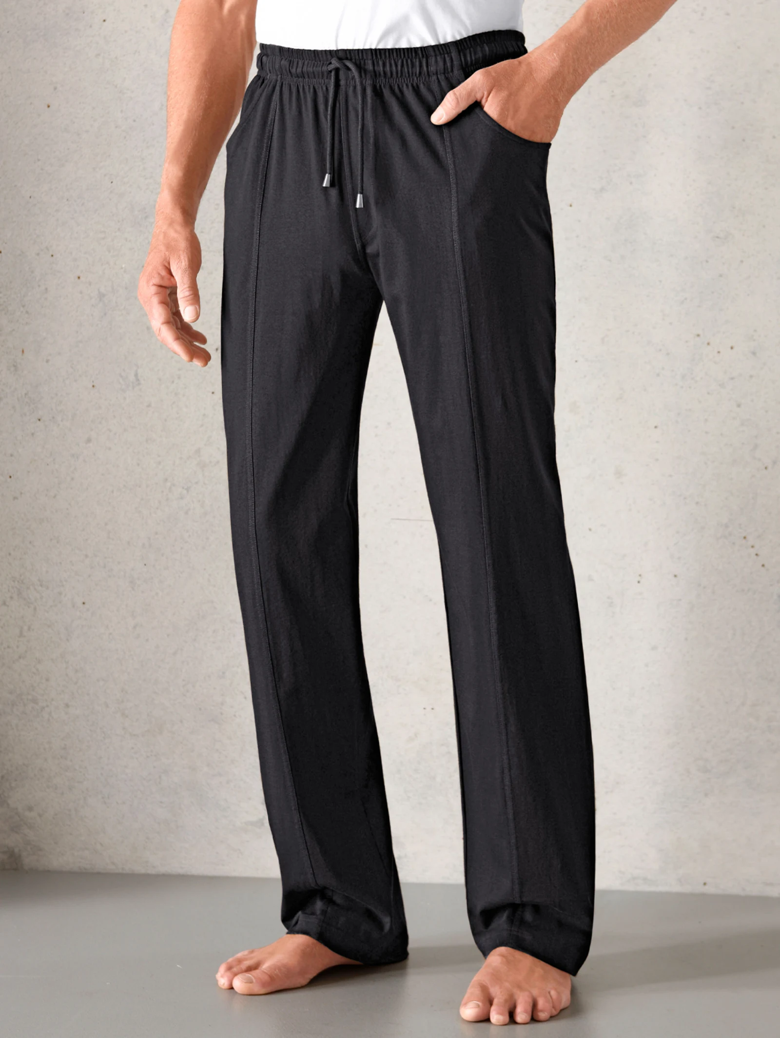 Schwarze Herren-Freizeithose mit elastischem Bund und Kordelzug, seitlichen Taschen, getragen mit weißem Shirt, barfuß auf grauem Boden. Schwarze Herren-Freizeithose mit elastischem Bund und Kordelzug, seitlichen Taschen, getragen mit weißem Shirt, barfuß auf grauem Boden.
