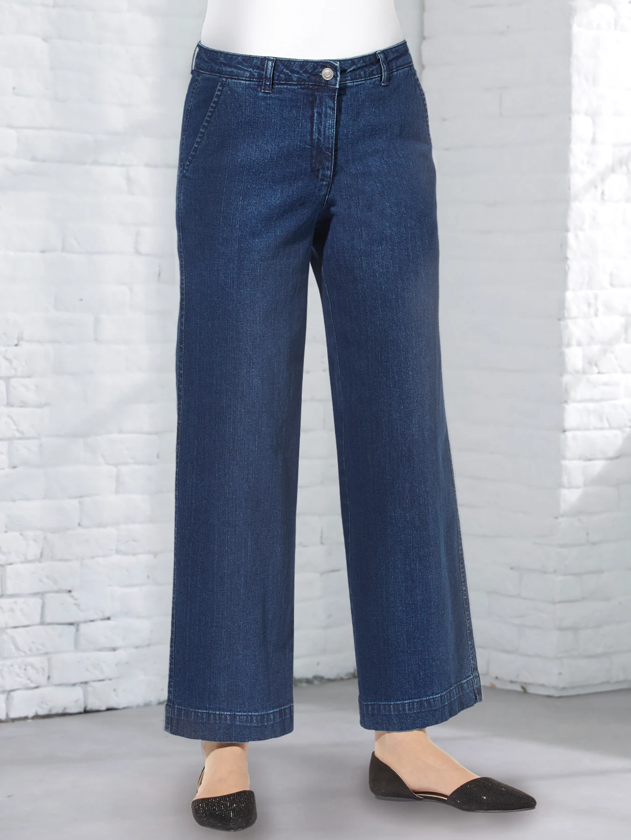 Dunkelblaue Jeans mit weitem Schnitt, getragen von einem Model, kombiniert mit schwarzen Ballerinas vor einer weißen Backsteinwand.