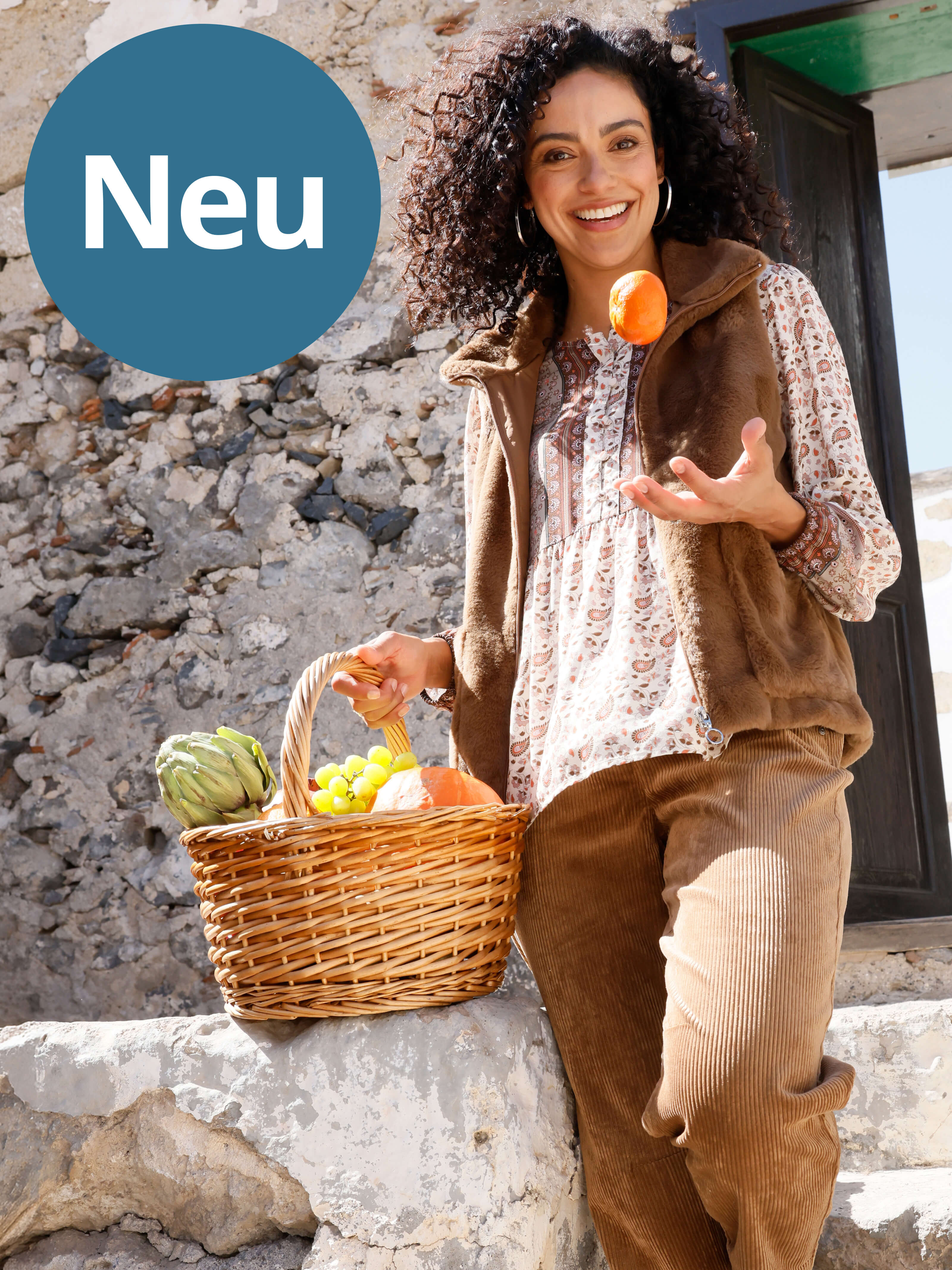 Ein Bild mit dem Text 'Neu'. Der Hintergrund zeigt eine lachende Frau mit lockigen Haaren, die einen Korb mit frischem Gemüse trägt und eine Tomate in der Hand hält. Sie steht vor einer alten Steinmauer und einer offenen Tür im Sonnenlicht.
