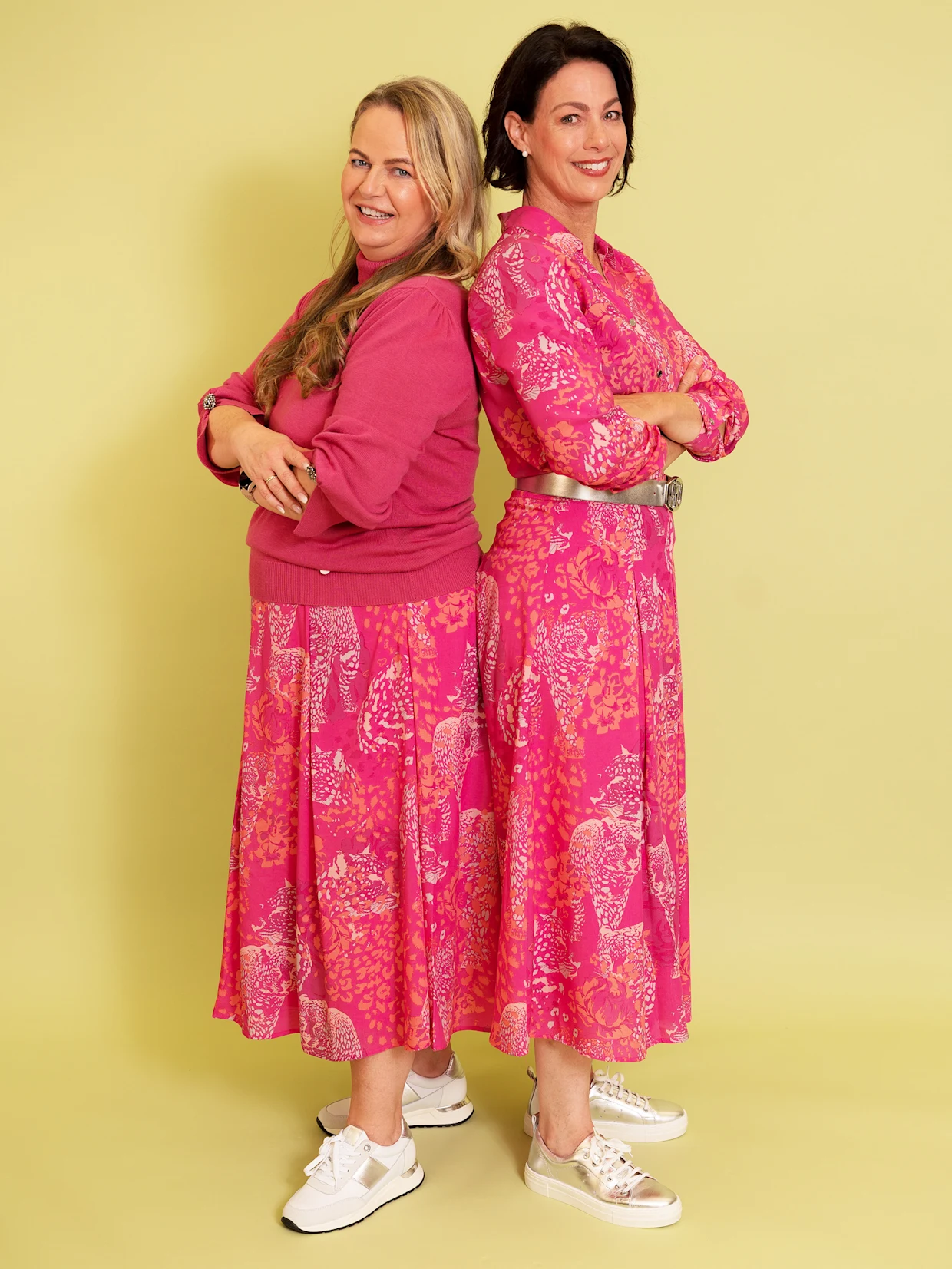 Zwei lächelnde Frauen stehen Rücken an Rücken vor einem hellgelben Hintergrund. Die Frau links hat blonde, wellige Haare und trägt einen pinken Pullover und einen pinken Rock mit abstraktem Muster, dazu weiße Sneaker mit goldenen Akzenten. Die Frau rechts hat kurze, dunkle Haare und trägt eine pink gemusterte Bluse und einen passenden Rock mit einem silbernen Gürtel, dazu silberne Sneaker. Beide Frauen haben die Arme verschränkt. Zwei lächelnde Frauen stehen Rücken an Rücken vor einem hellgelben Hintergrund. Die Frau links hat blonde, wellige Haare und trägt einen pinken Pullover und einen pinken Rock mit abstraktem Muster, dazu weiße Sneaker mit goldenen Akzenten. Die Frau rechts hat kurze, dunkle Haare und trägt eine pink gemusterte Bluse und einen passenden Rock mit einem silbernen Gürtel, dazu silberne Sneaker. Beide Frauen haben die Arme verschränkt.