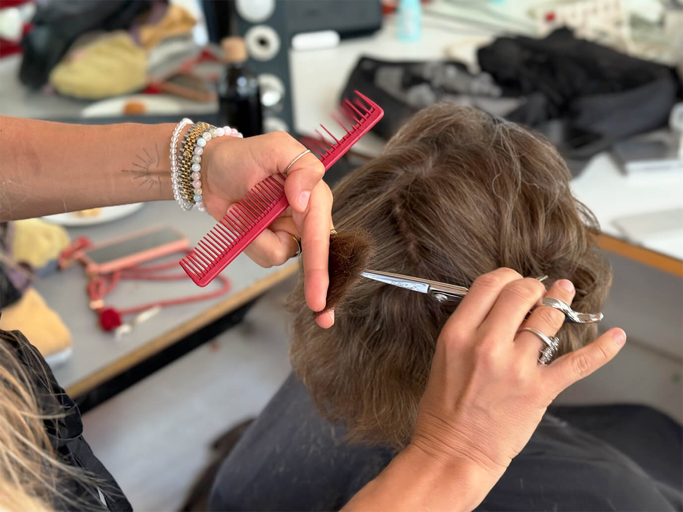 Frau sitzt mit dem Rücken zur Kamera. Friseurin schneidet ihr braunes Haar mit Schere und rotem Kamm, Nahaufnahme der Hände und Kopf, im Hintergrund Friseurutensilien unscharf.