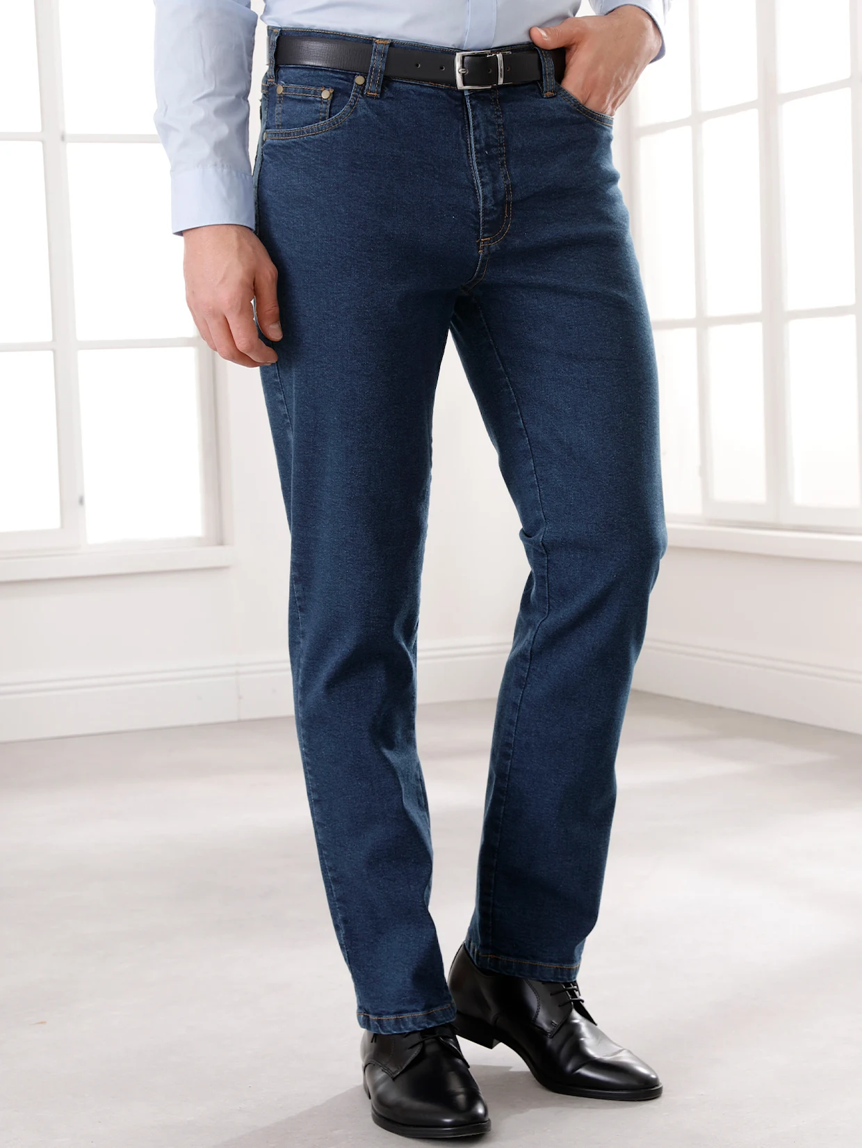 Mann trägt dunkelblaue Jeans, hellblaues Hemd, schwarzen Ledergürtel und schwarze Schuhe, stehend vor einem hellen Fenster. Mann trägt dunkelblaue Jeans, hellblaues Hemd, schwarzen Ledergürtel und schwarze Schuhe, stehend vor einem hellen Fenster.
