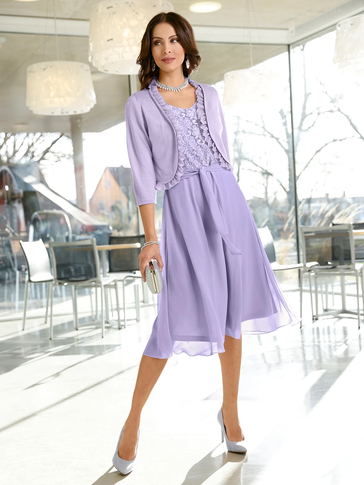 Frau in eleganter lila Abendgarderobe mit Spitzenoberteil, passender Strickjacke und A-Linien-Rock, kombiniert mit lilafarbenen High Heels und silberner Clutch, stehend in modernem, lichtdurchflutetem Raum.