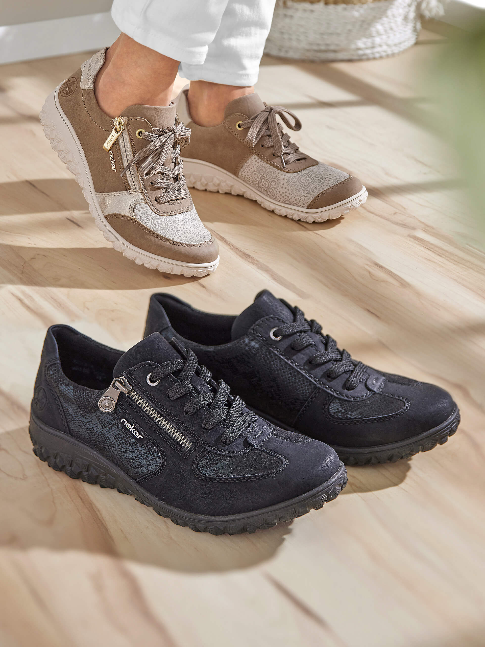 Eine Frau trägt ein Paar beige Sneaker, daneben steht ein Paar schwarze Sneaker auf einem hellen Holzboden.