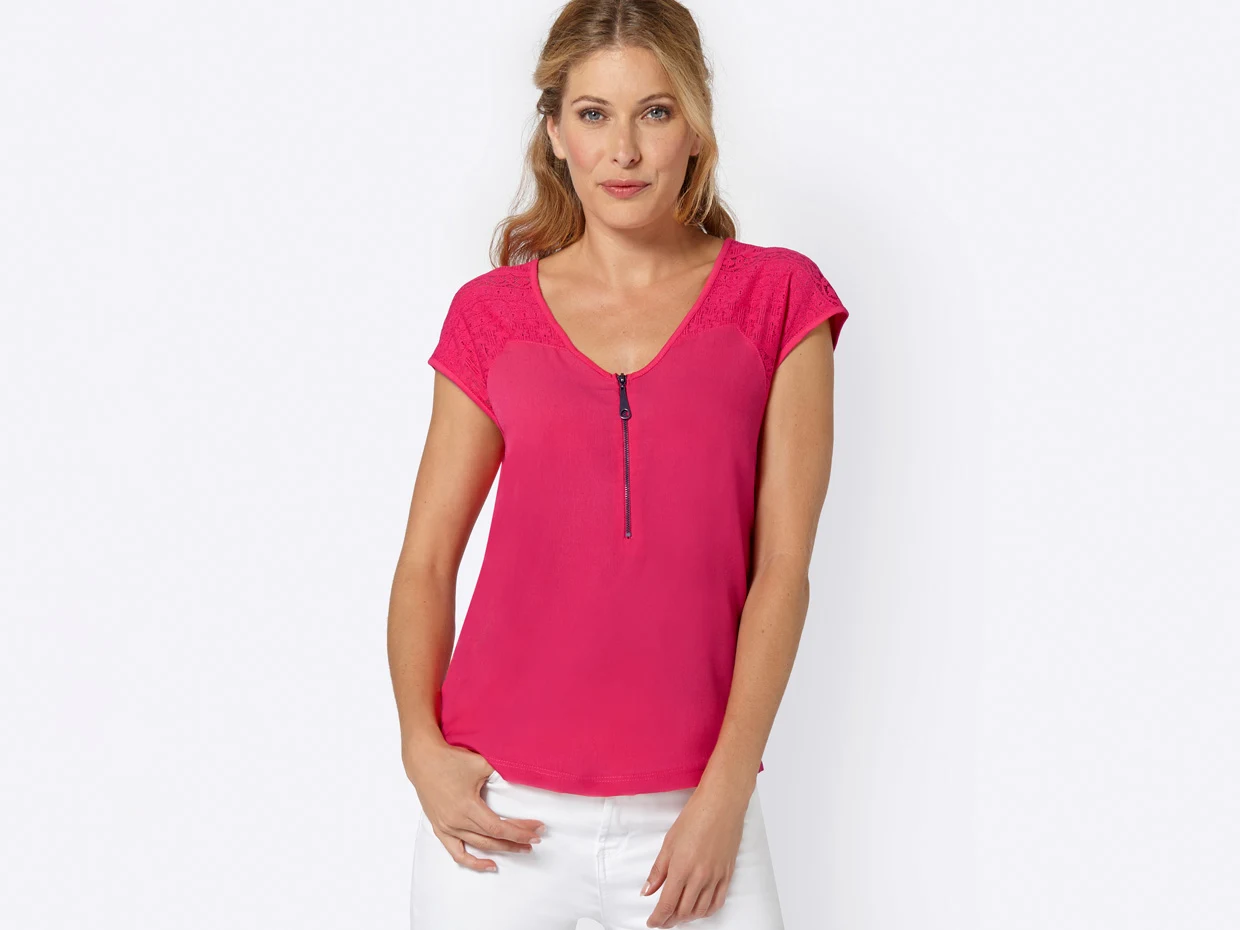 Damen-Shirt in kräftigem Pink mit V-Ausschnitt, Reißverschluss-Detail und kurzen Ärmeln aus Spitze, kombiniert mit weißer Hose.