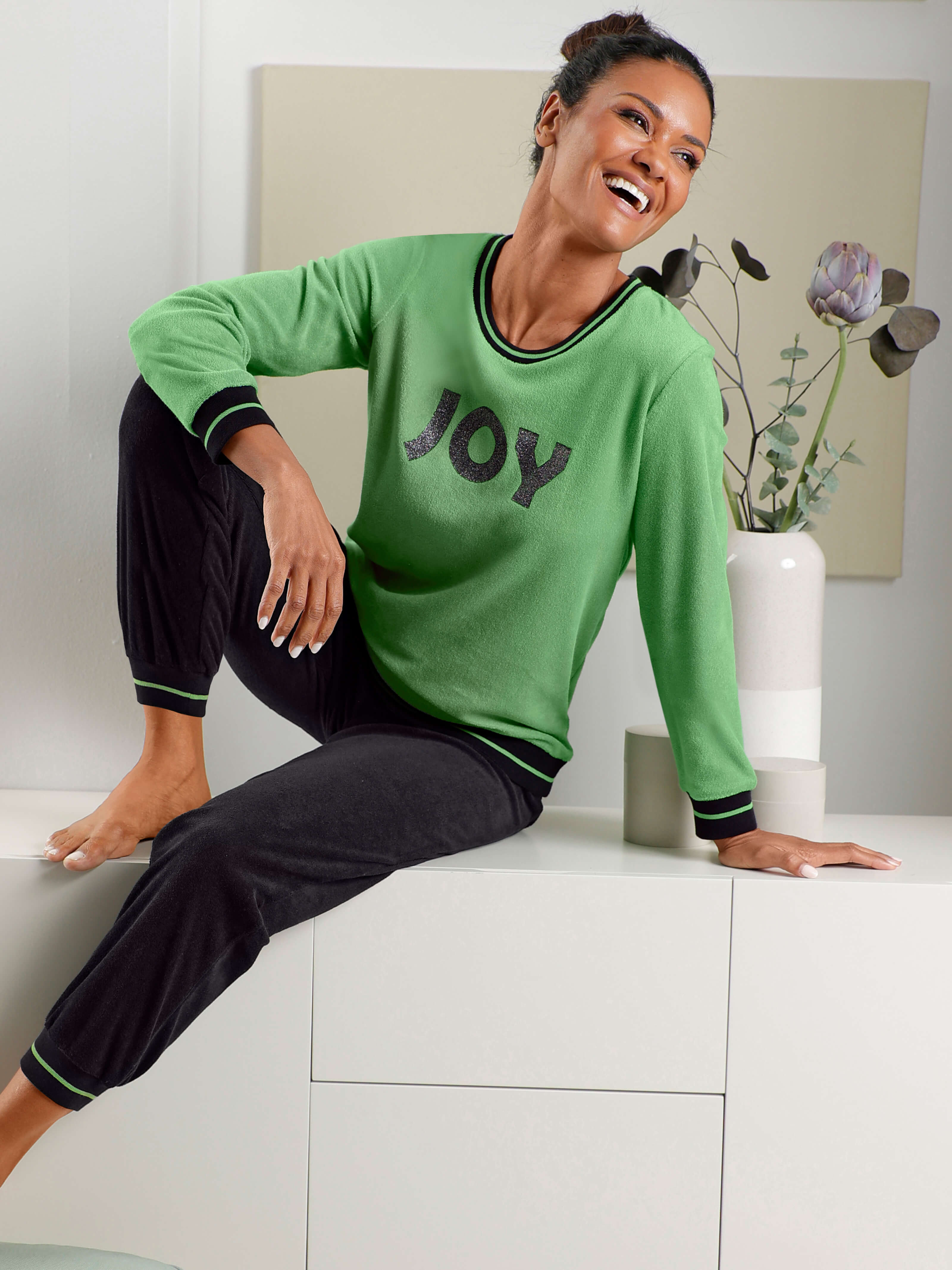 Ein Model trägt ein grünes Oberteil mit Aufschrift 'JOY' und eine schwarze Hose in einem hellen Raum.