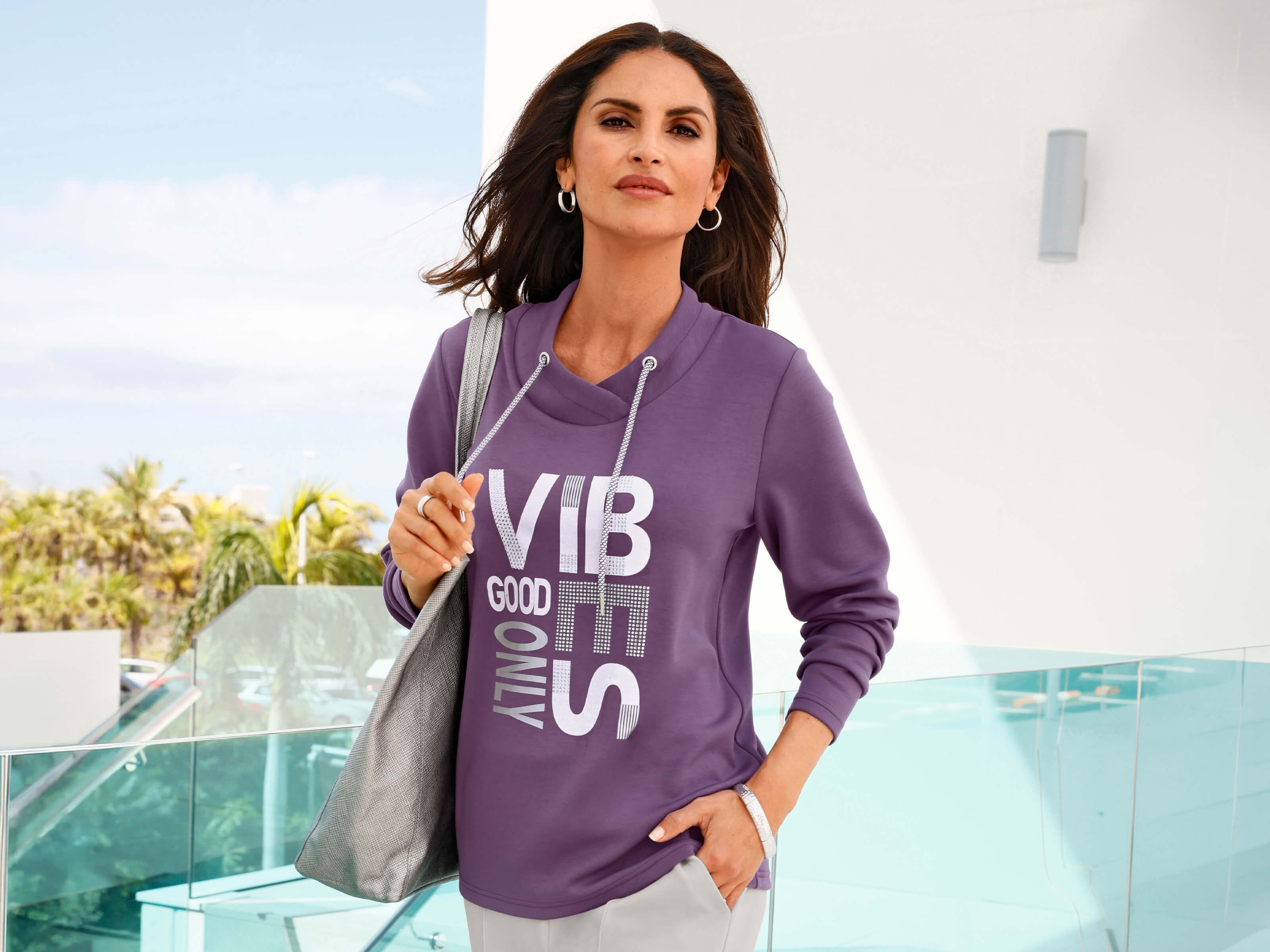 Ein Model trägt einen violetten Pullover, helle Hose und silberne Schultertasche an einer Glastreppe.