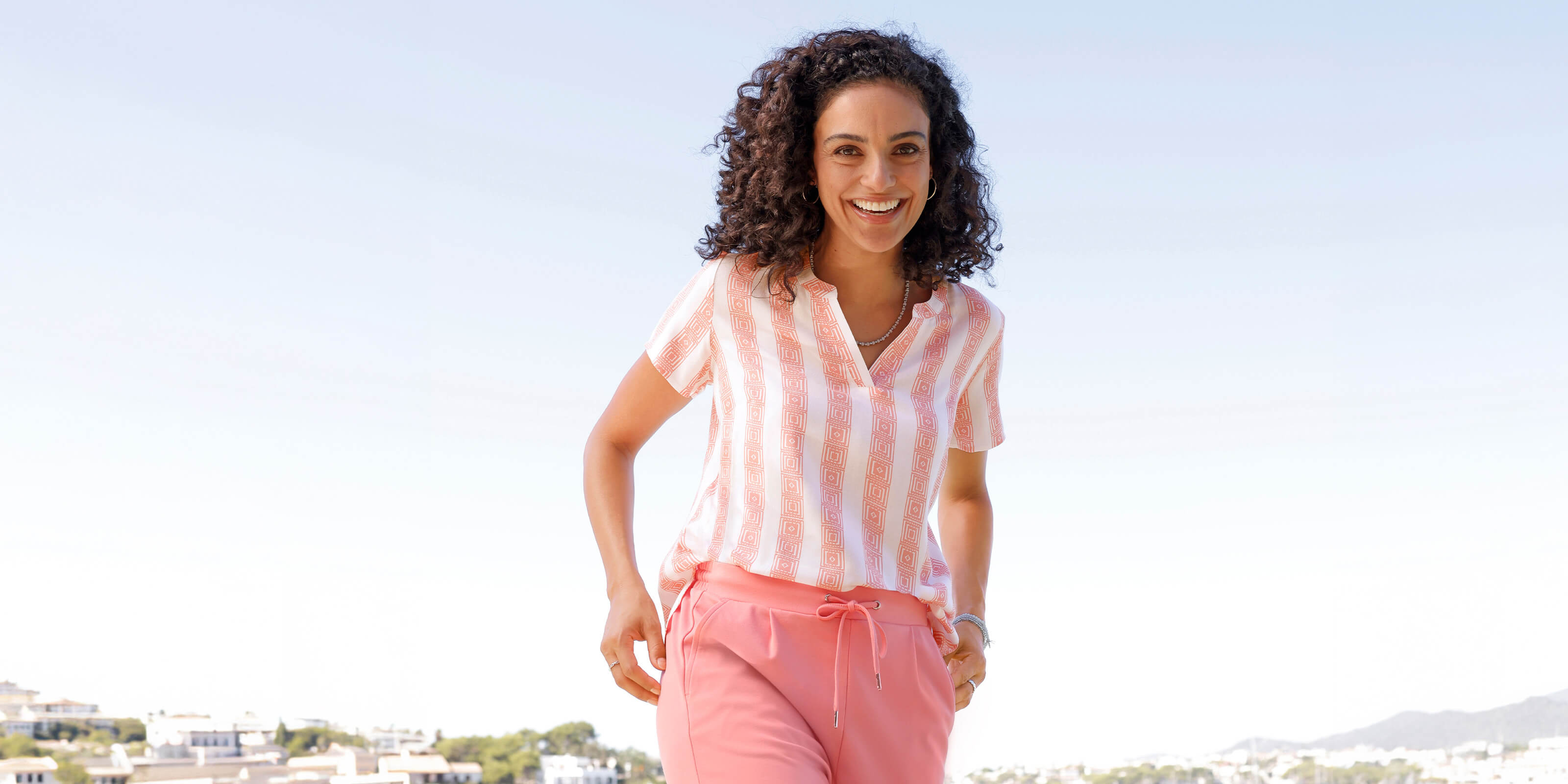 Ein Model trägt eine kurzärmelige Bluse mit rosa Streifen und eine passende rosa Hose mit Kordelzug, steht am Wasser.