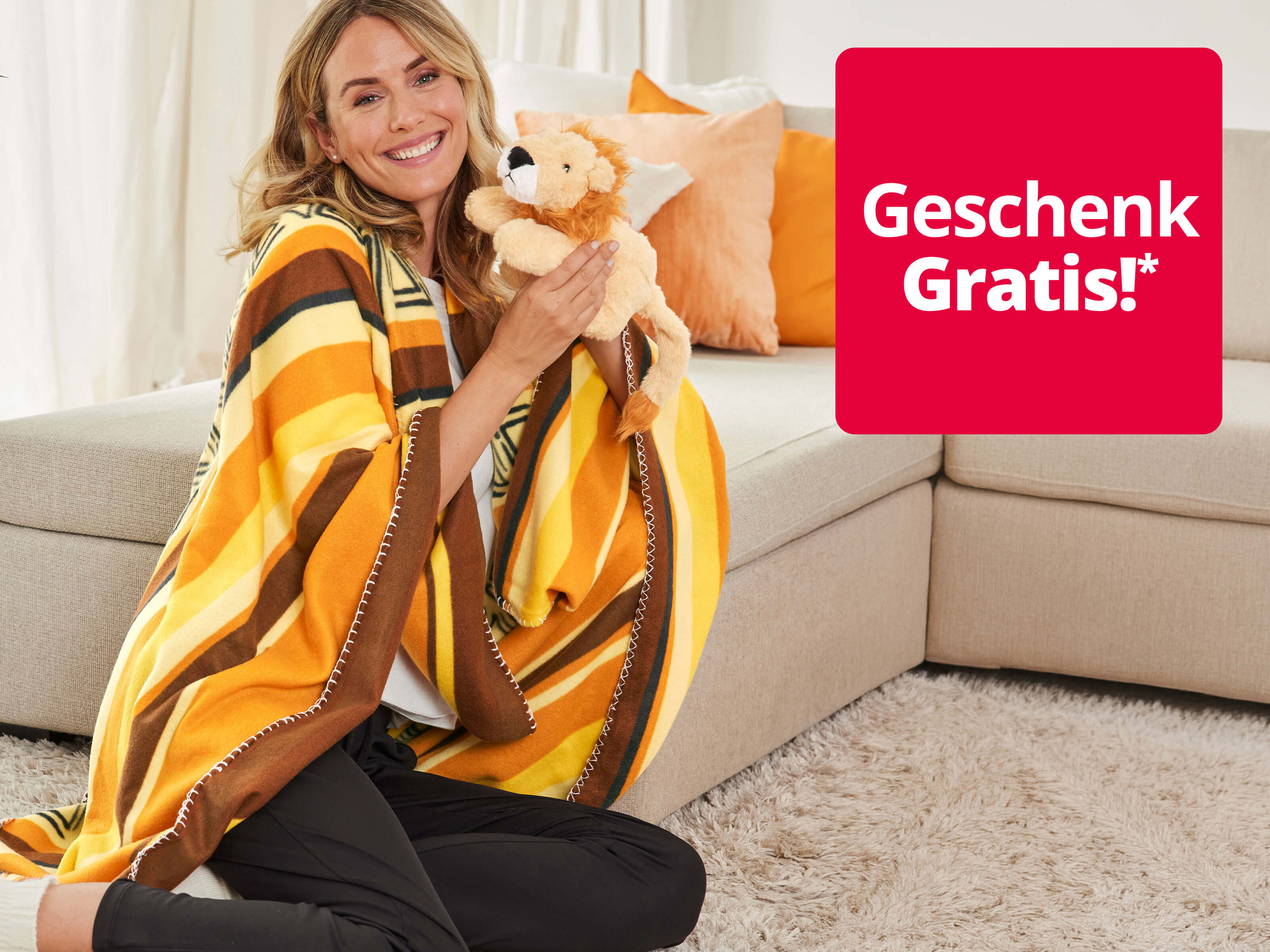 Ein Bild mit dem Text 'Geschenk Gratis!*'. Der Hintergrund zeigt eine lächelnde Frau in einer gelb-braunen Decke, die neben einem beigen Sofa sitzt und ein Plüsch-Löwenkuscheltier hält.