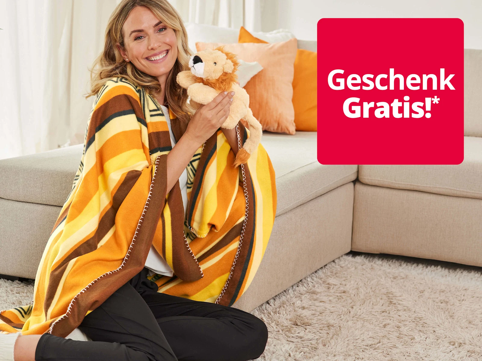Ein Bild mit dem Text 'Geschenk Gratis!*'. Der Hintergrund zeigt eine lächelnde Frau in einer gelb-braunen Decke, die neben einem beigen Sofa sitzt und ein Plüsch-Löwenkuscheltier hält. Ein Bild mit dem Text 'Geschenk Gratis!*'. Der Hintergrund zeigt eine lächelnde Frau in einer gelb-braunen Decke, die neben einem beigen Sofa sitzt und ein Plüsch-Löwenkuscheltier hält.