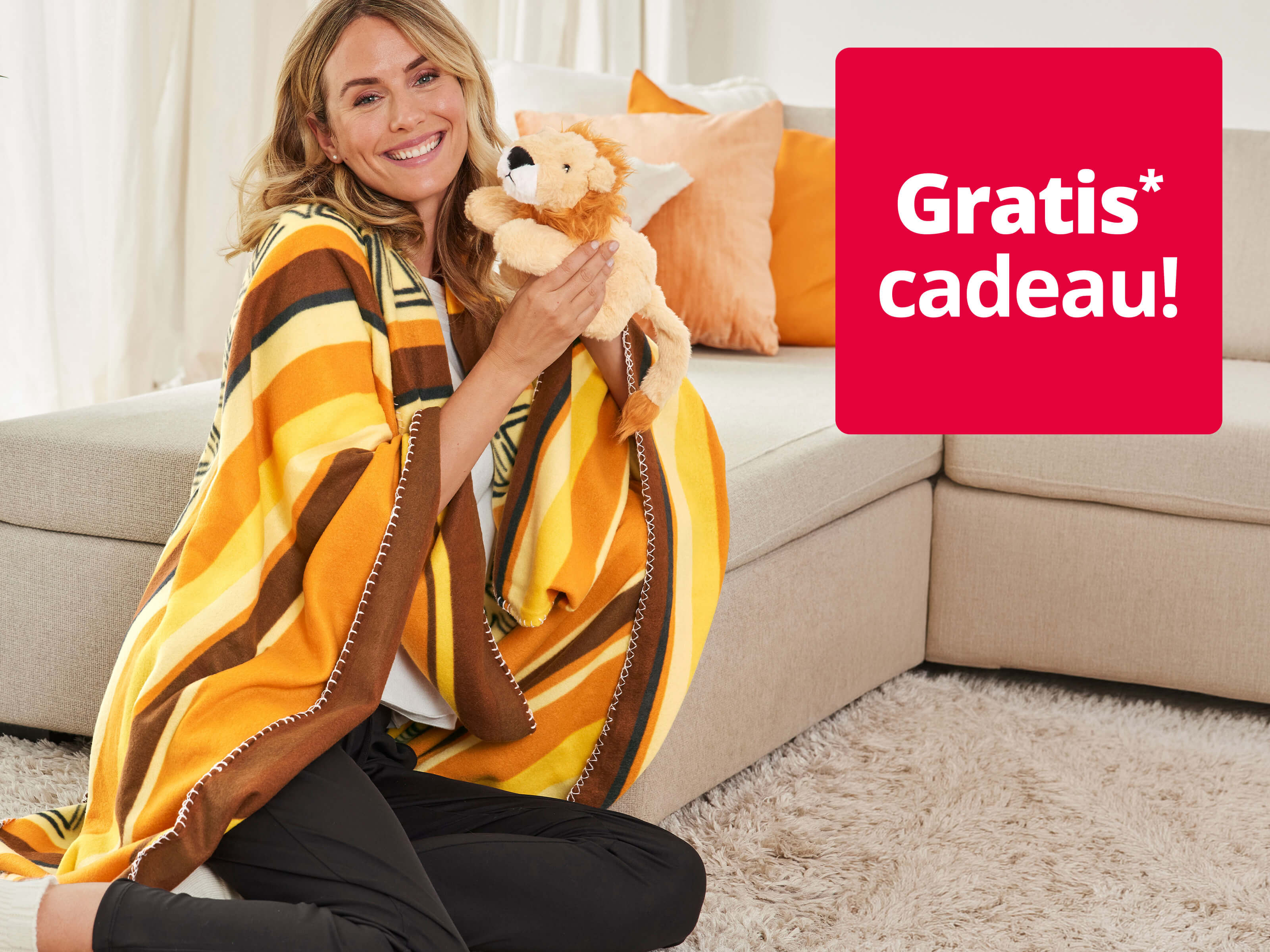 Een afbeelding met de tekst 'Gratis* cadeau!'. Op de achtergrond is een glimlachende vrouw te zien in een geelbruine deken, die naast een beige bank zit en een pluchen leeuw vasthoudt.