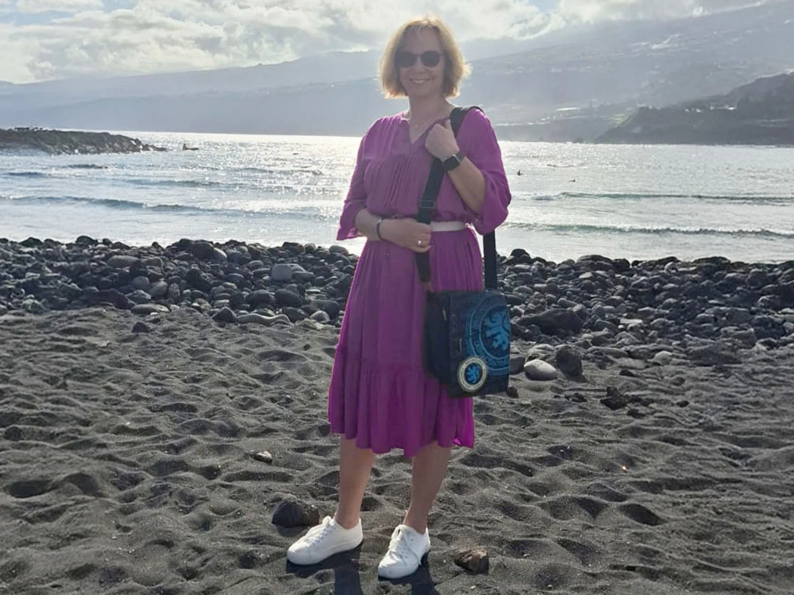 Eine Frau steht am Strand auf dunklem Sand, trägt ein lila Kleid, weiße Schuhe und hält eine Tasche. Im Hintergrund ist das Meer sichtbar. Eine Frau steht am Strand auf dunklem Sand, trägt ein lila Kleid, weiße Schuhe und hält eine Tasche. Im Hintergrund ist das Meer sichtbar.