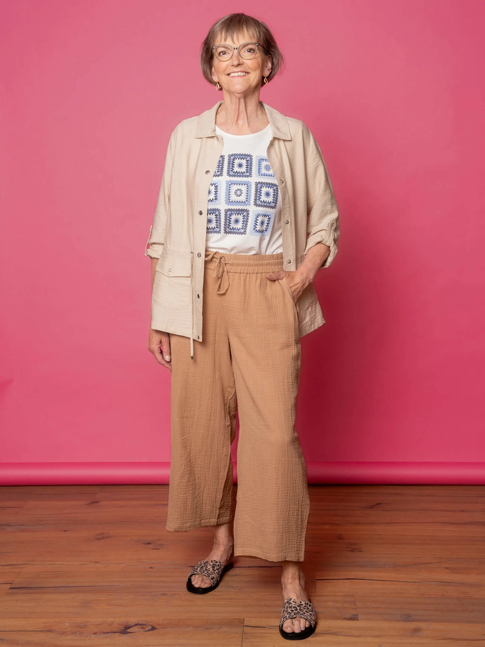 Eine Frau trägt eine beige Jacke über einem weißen T-Shirt mit blauem Muster und einer braunen, weiten Hose. Sie trägt Sandalen mit Leopardenmuster. Der Hintergrund ist pink.